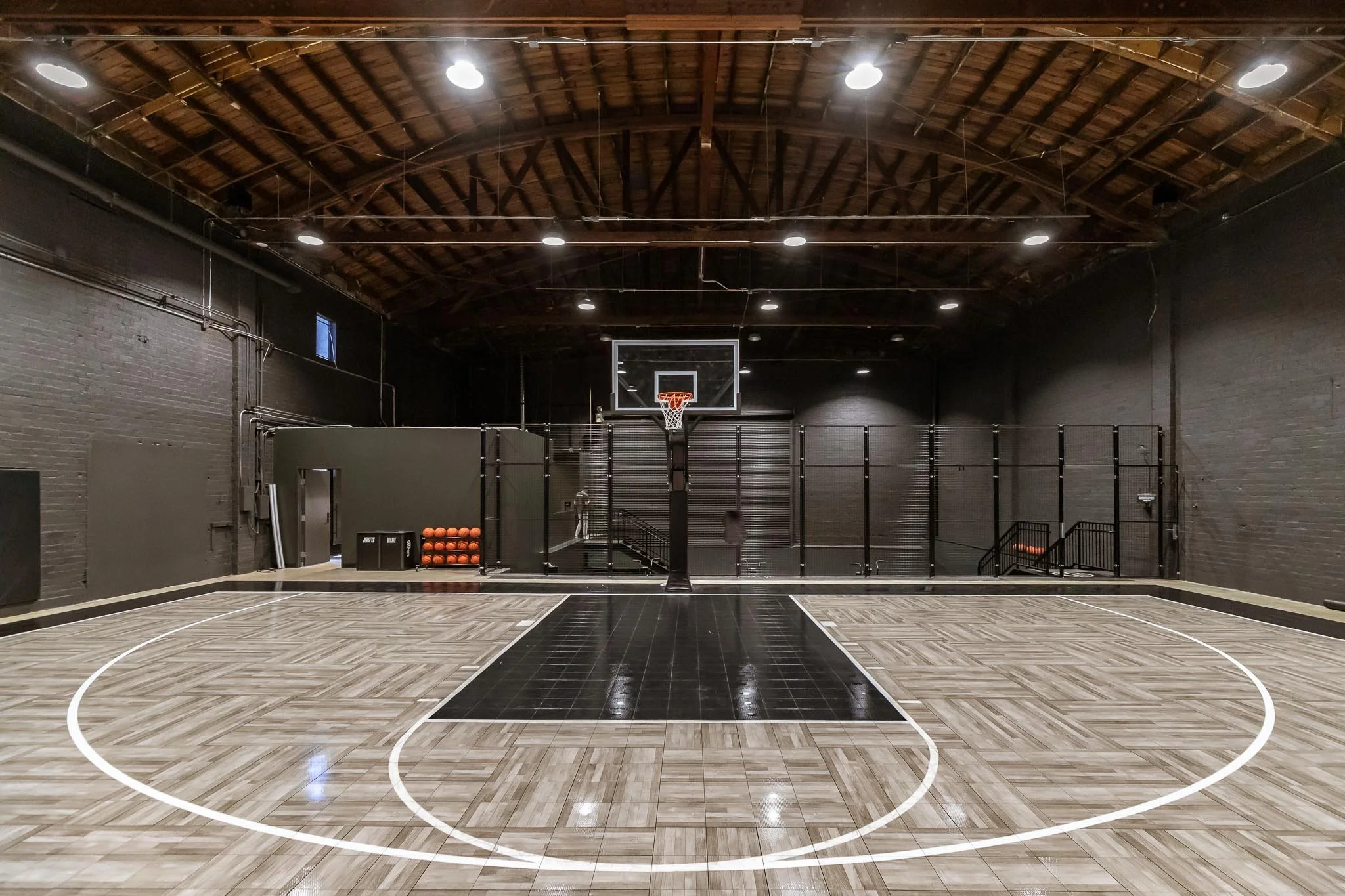 Empty indoor basketball court with black and wood-colored flooring, basketball hoop in center, black walls, and ceiling with wooden beams and lights.