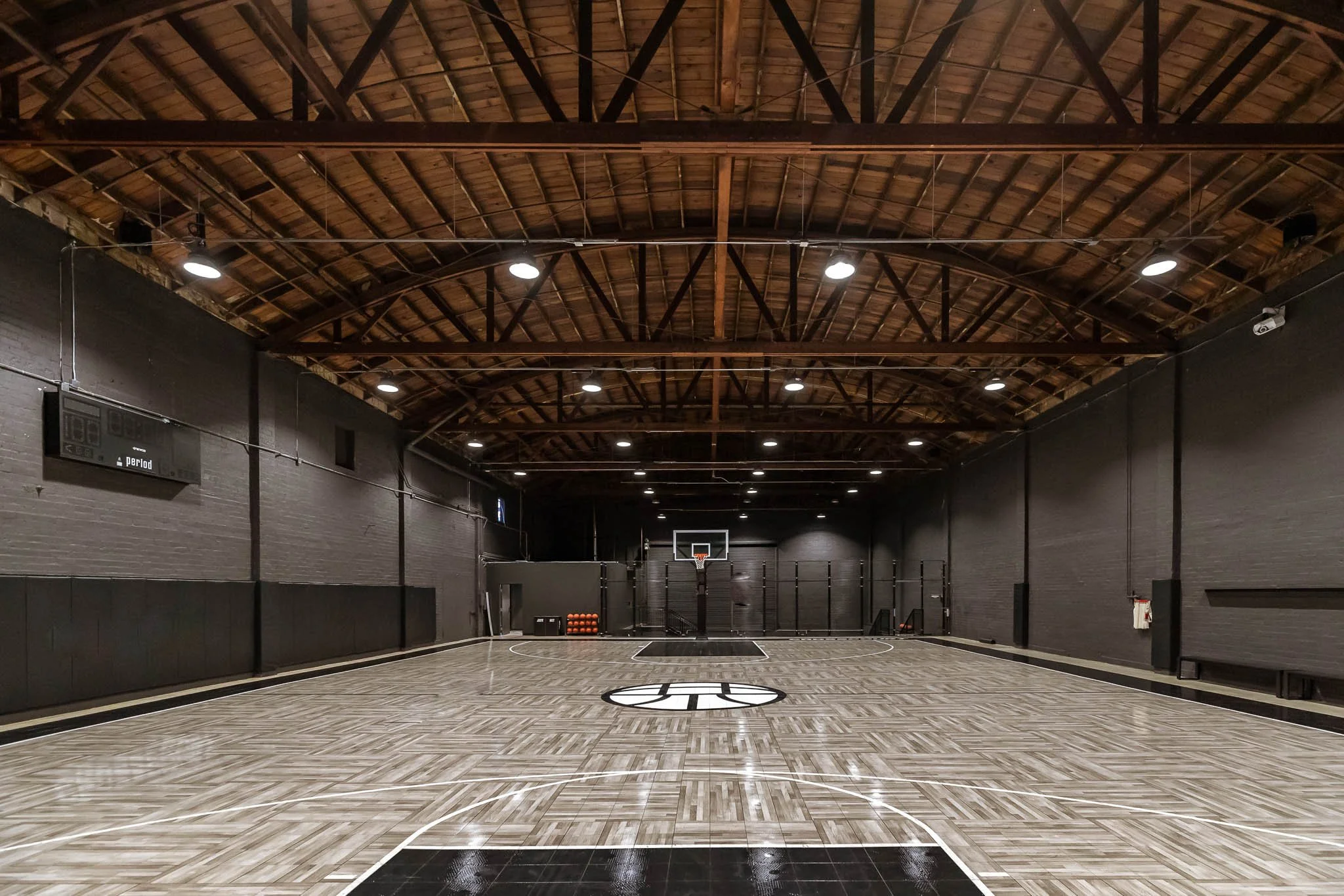 Empty indoor basketball court with hardwood flooring, basketball hoop, and black walls, under a wooden ceiling with lights.