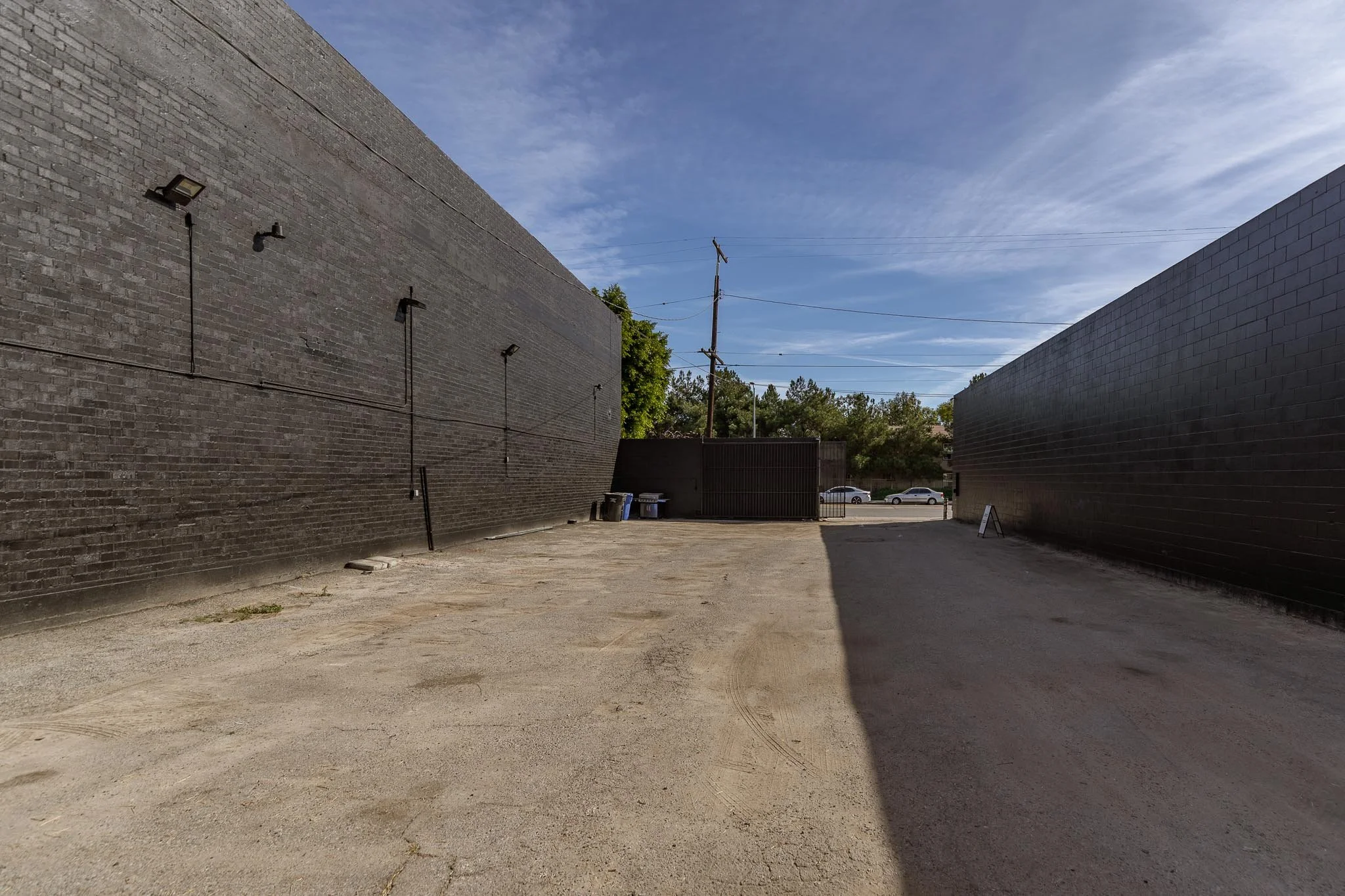 An empty dirt lot between two tall brick buildings, with a dark gate and a few trash bins at the end, under a partly cloudy sky.