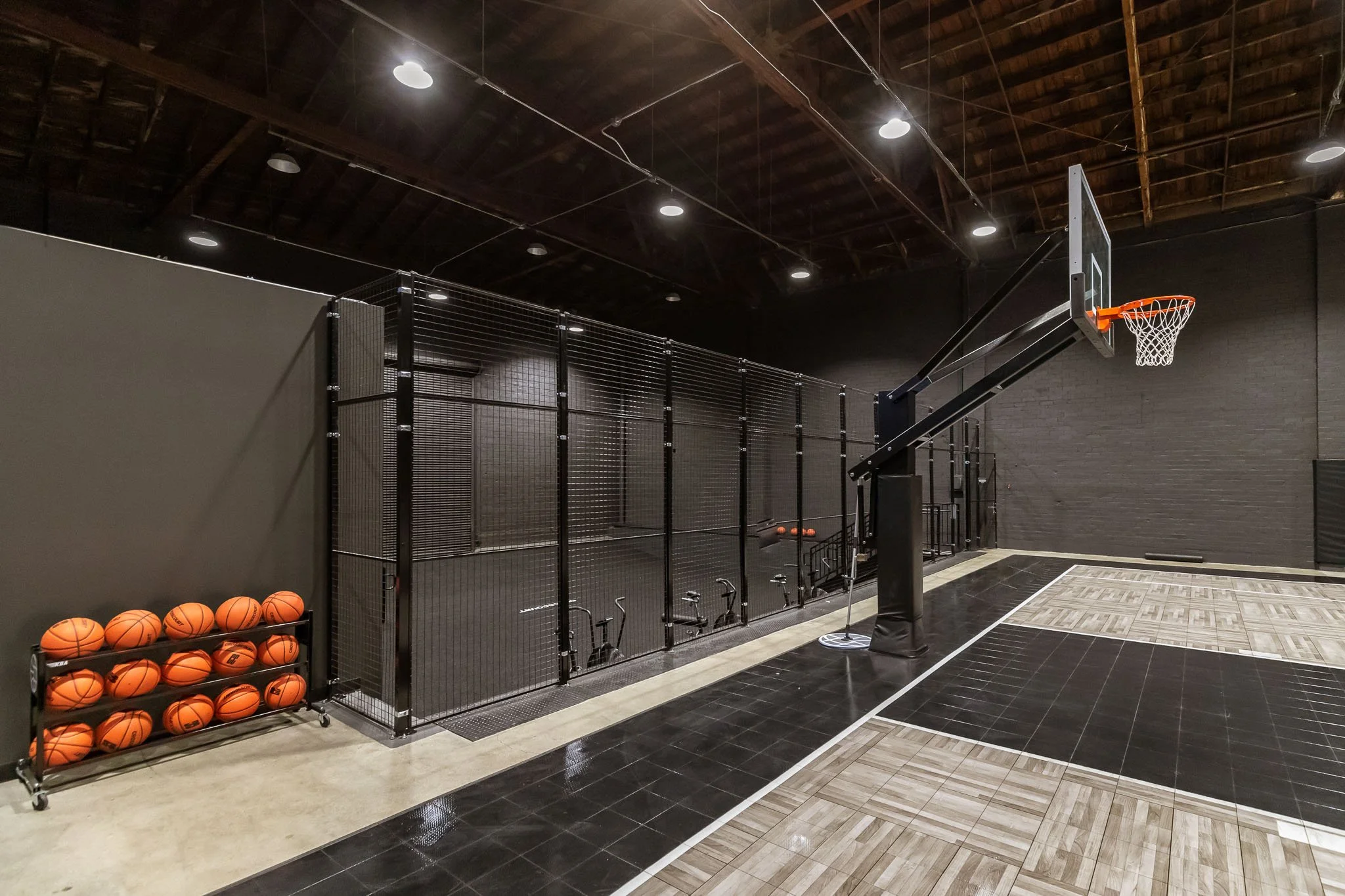 Indoor basketball court with a hoop, a rack of basketballs, and a fenced-off area, with dark walls and ceiling, and mixed tile and wooden flooring.