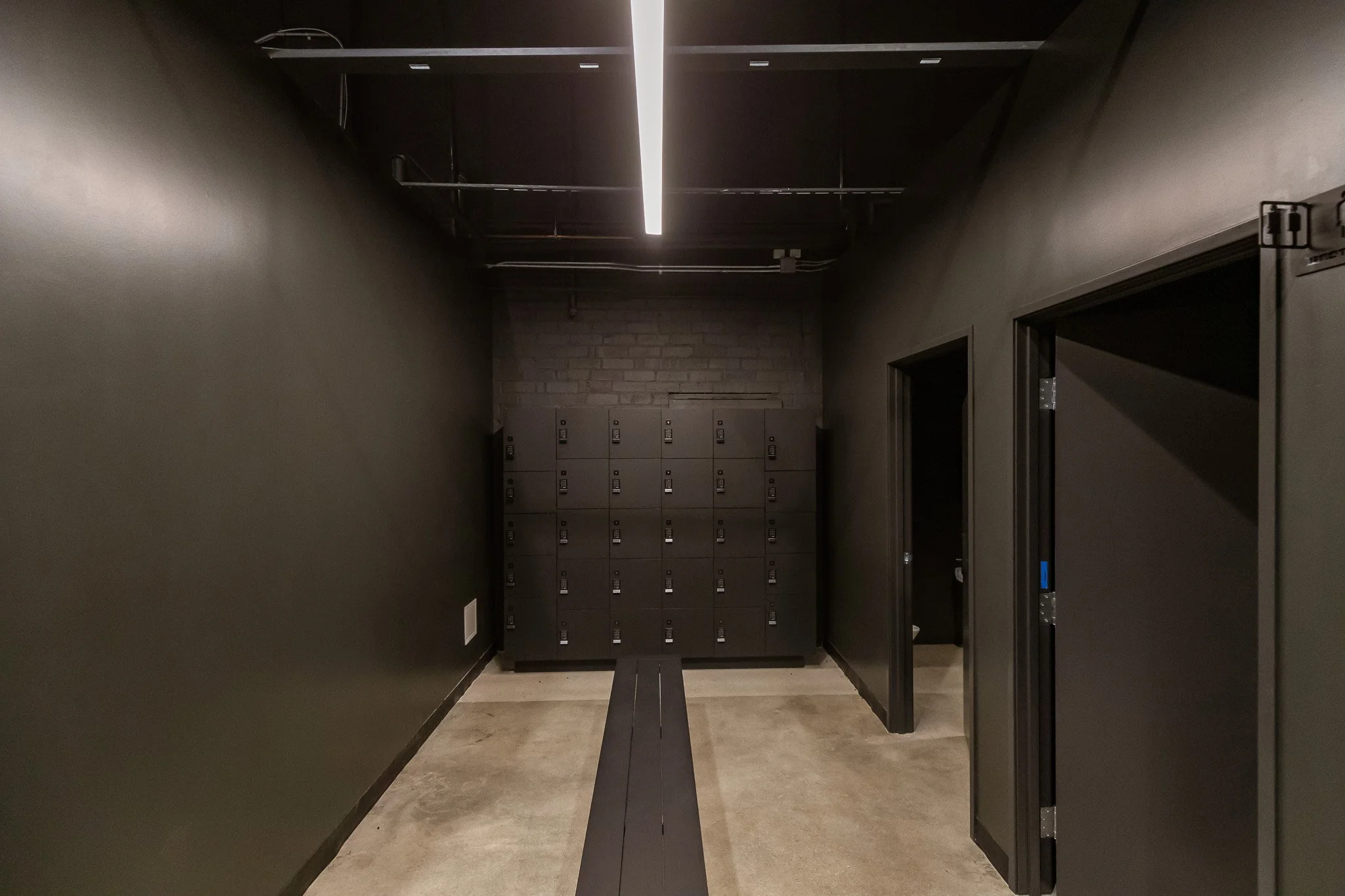 A hallway with dark walls and ceiling, fluorescent lighting, and several black lockers at the end.