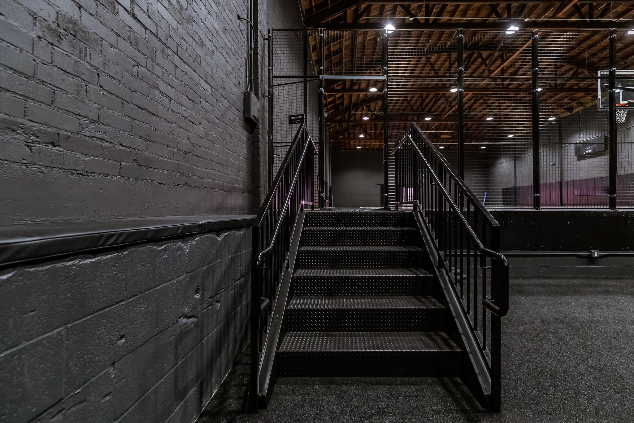 Indoor skate park with metal stairs and black grid fence, brick wall on left, illuminated ceiling, and basketball hoop in the background.