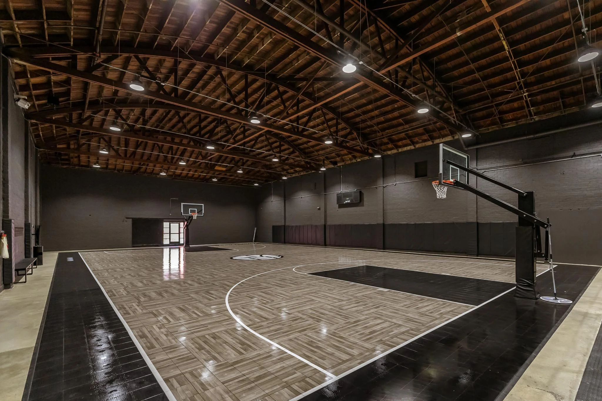 Empty indoor basketball court with wooden flooring, black padded walls, and a high ceiling with exposed wood beams. There are two basketball hoops, one at each end of the court, and bright ceiling lights.