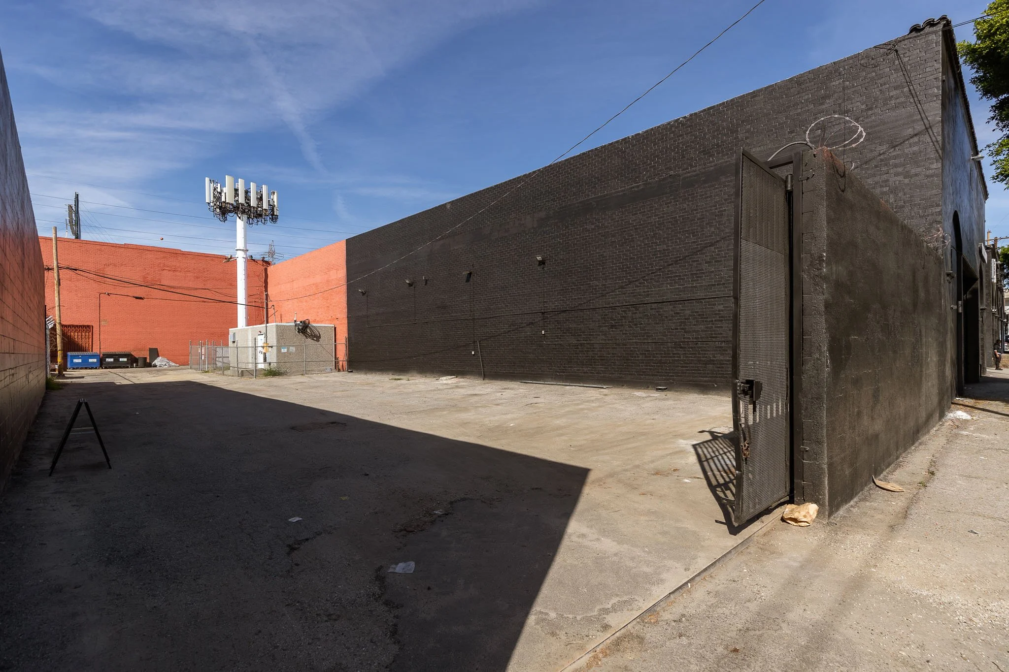 An empty outdoor lot surrounded by black and red brick walls, with a tall cell phone tower, electrical wires, and a blue sky with clouds.