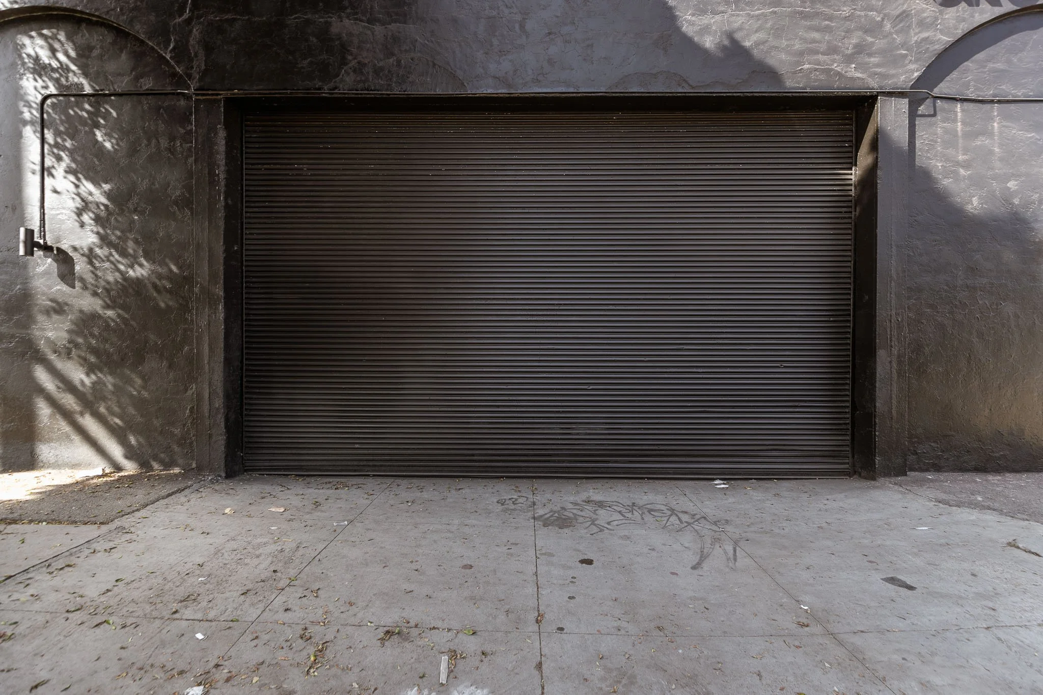 Black roll-up garage door on a concrete sidewalk with shadows and some graffiti