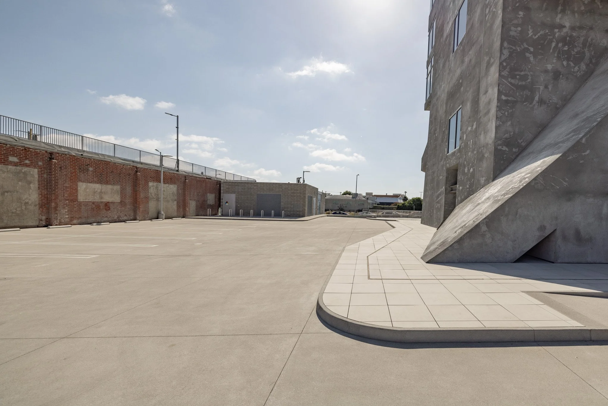 Empty parking lot next to a modern concrete building with a slanted wall, under a partly cloudy sky.