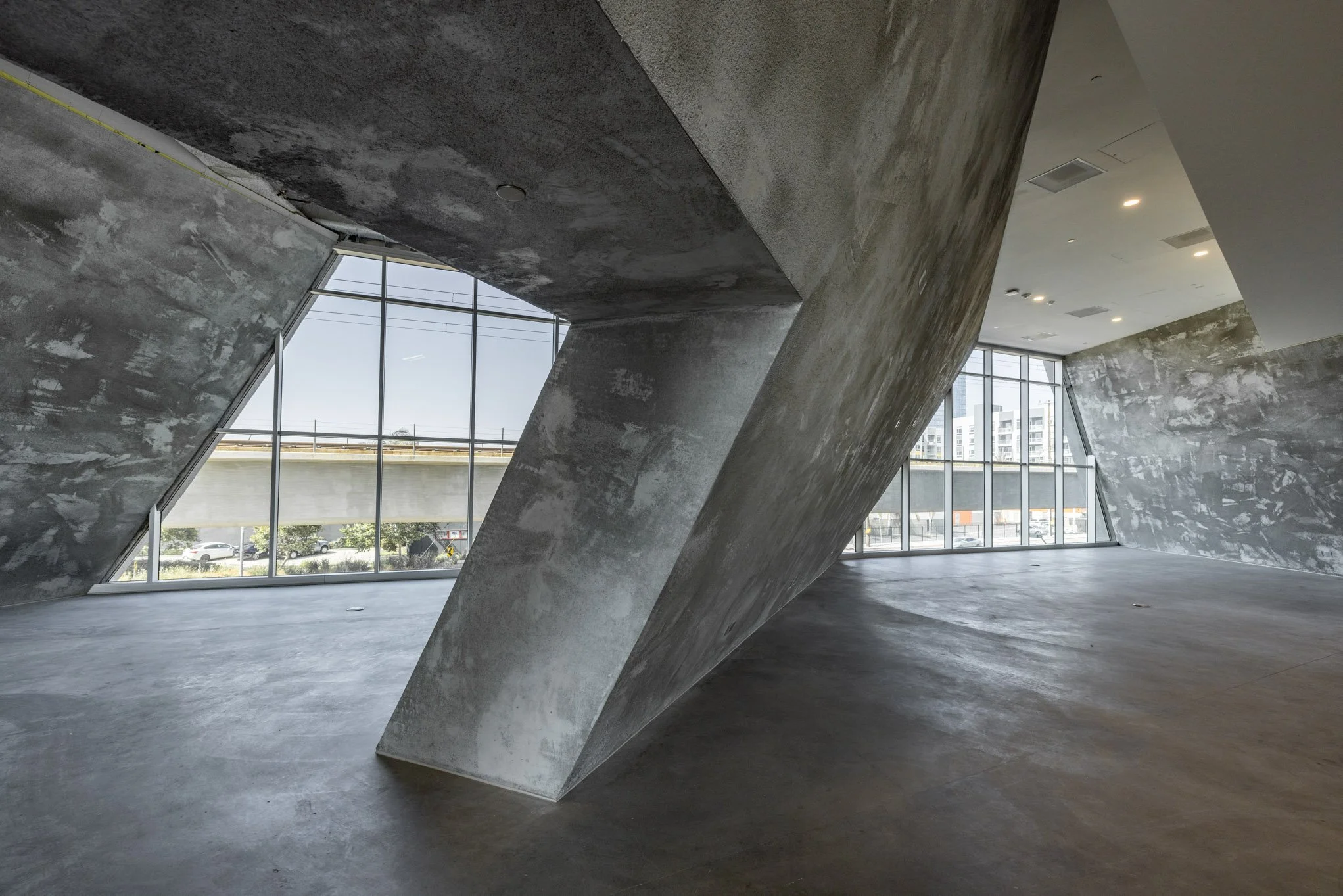 Interior space with modern, unfinished concrete walls and a large angular concrete pillar, featuring floor-to-ceiling windows showing an urban view.