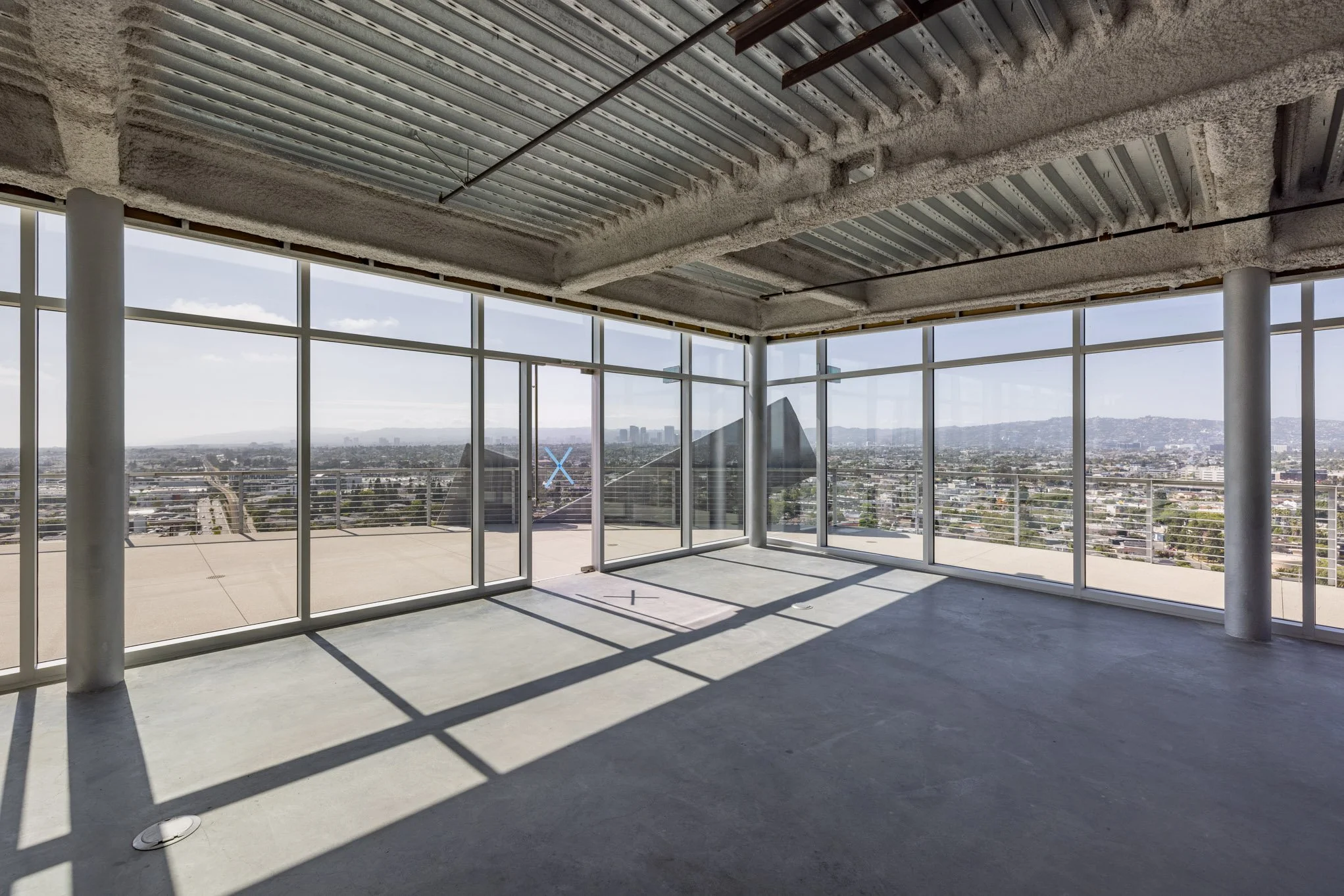Empty unfinished room with large glass windows showing a city skyline and mountains, concrete floor and ceiling, metal supports, and construction markings on the window