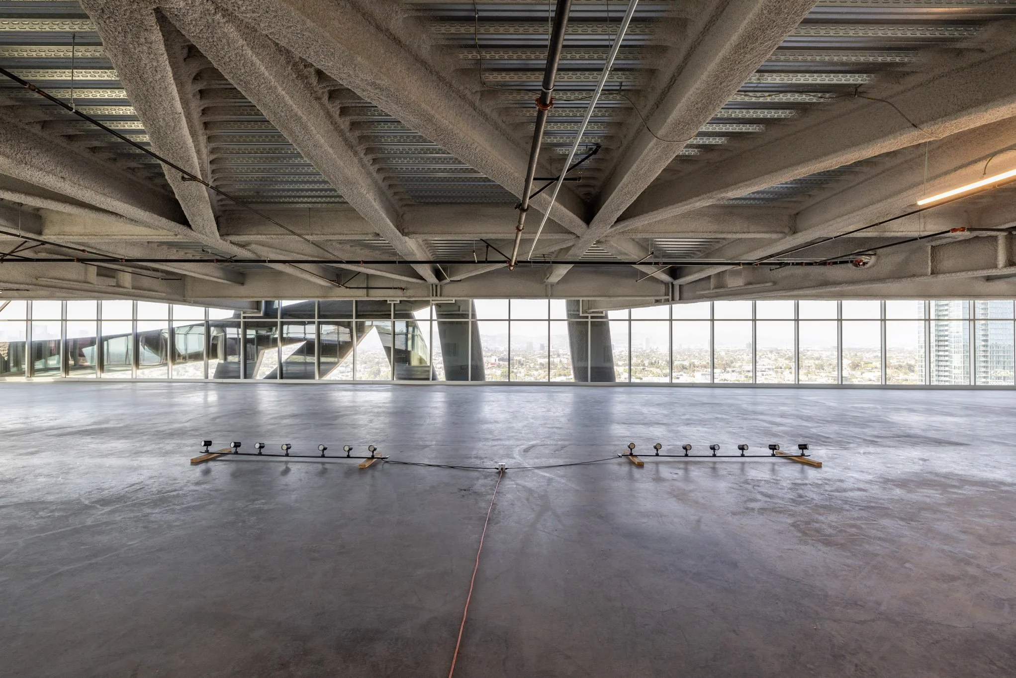 Empty indoor space under construction or renovation with large glass windows and cityscape view, construction lights on the floor, and exposed ceiling details.