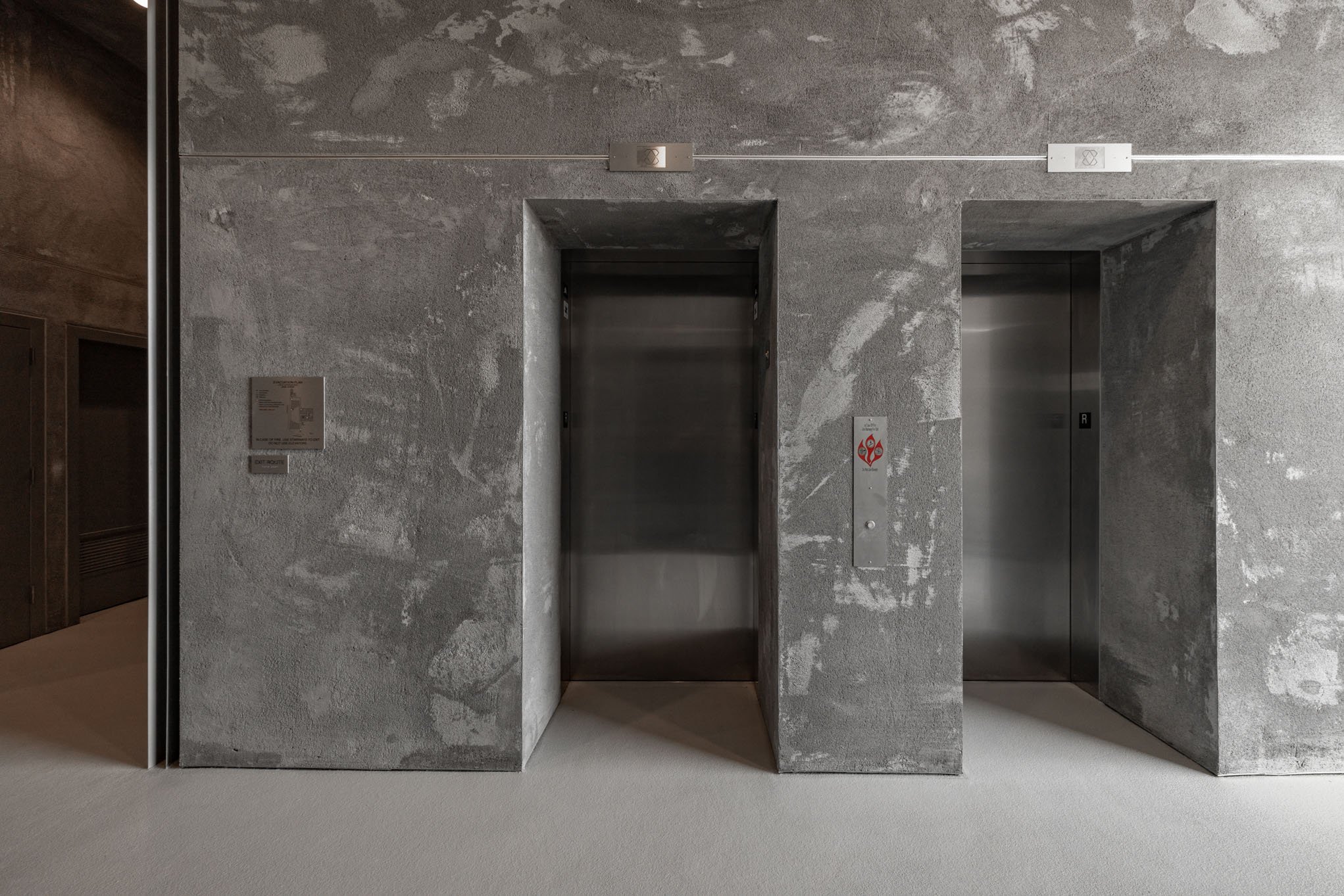 Two elevator doors set in a gray, textured concrete wall with minimal signage.