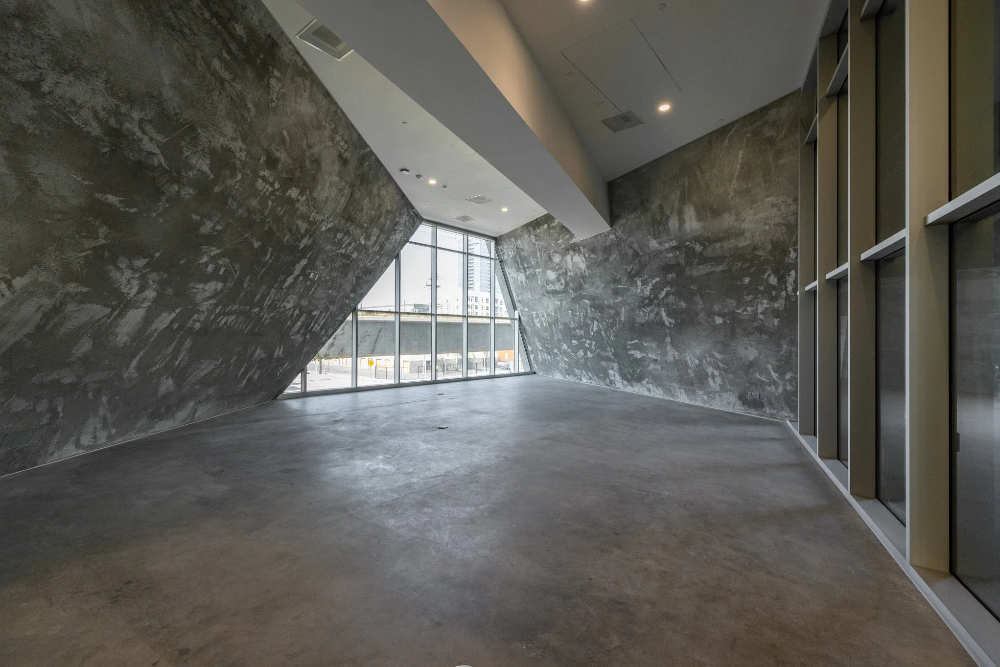 Empty modern interior space with concrete walls, a polished concrete floor, large floor-to-ceiling windows, and ceiling lights.