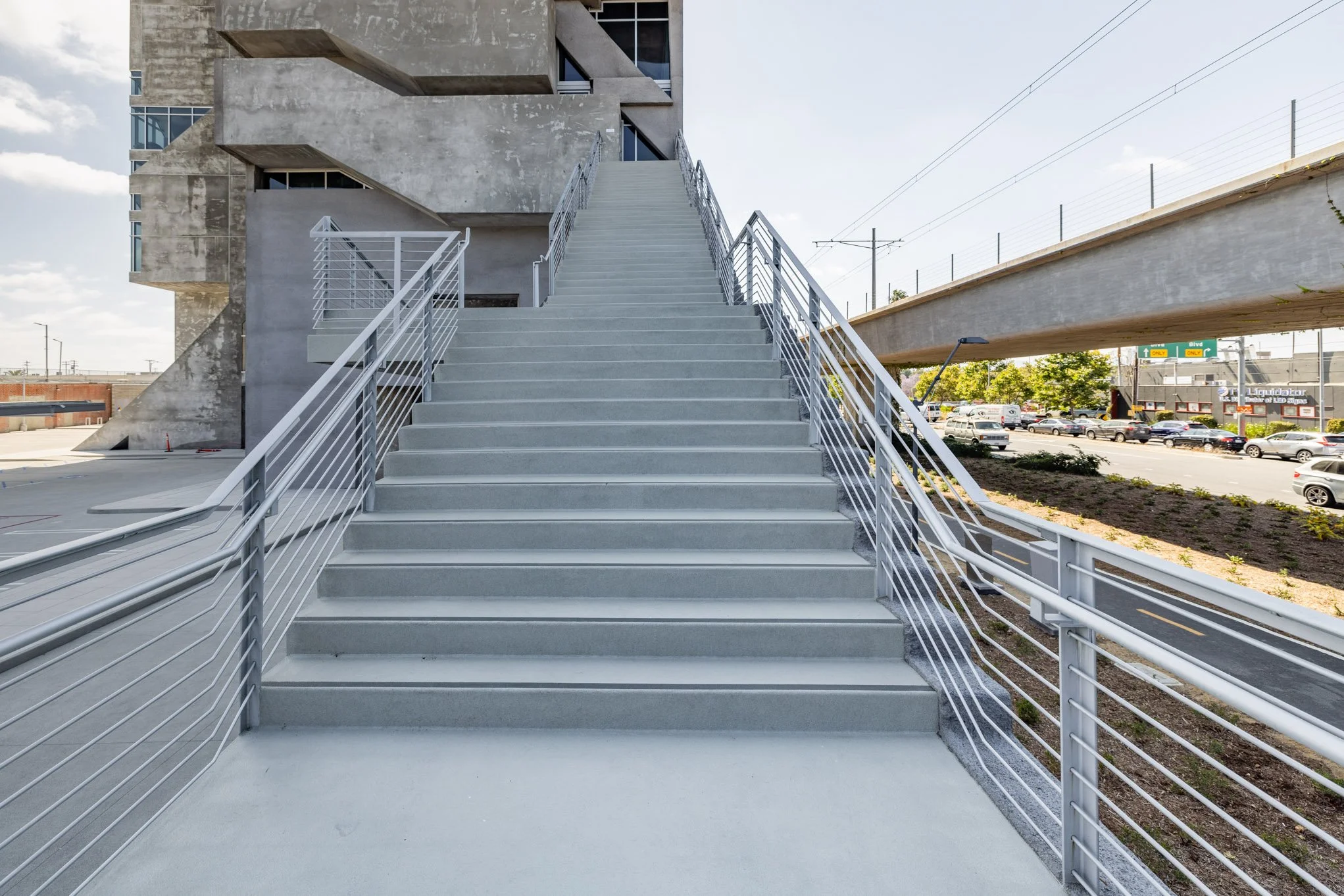 Staircase leading to a building with an industrial style, metal railings, concrete steps, and an elevated walkway beside a highway with cars in the background.
