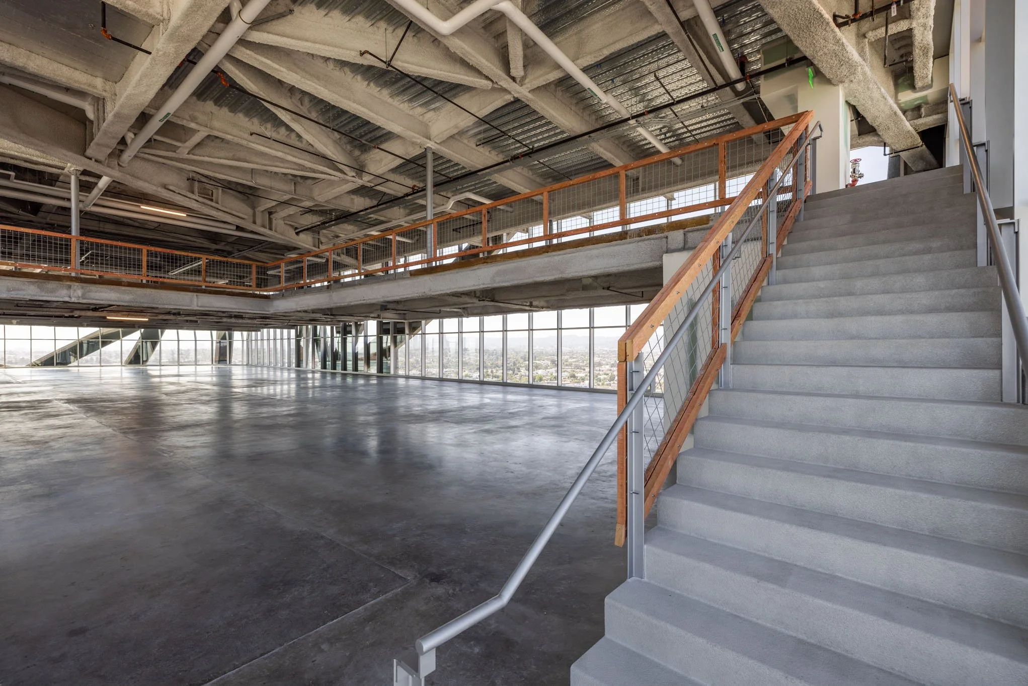 Interior view of a modern, unfinished building with concrete floors, exposed ceilings, large windows, and a staircase with metal railing and wooden handrail.