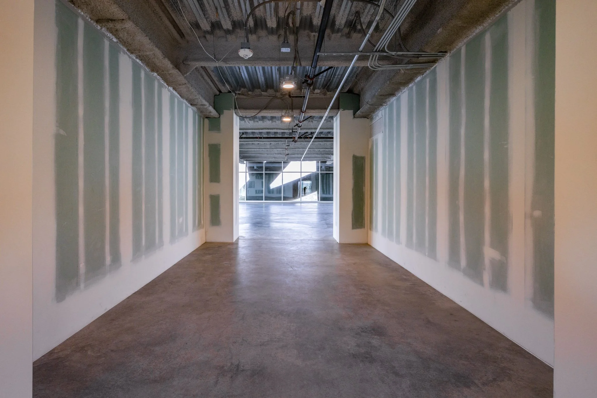 Construction site hallway with unfinished drywall on walls and exposed ceiling pipes, leading to a large window at the end.