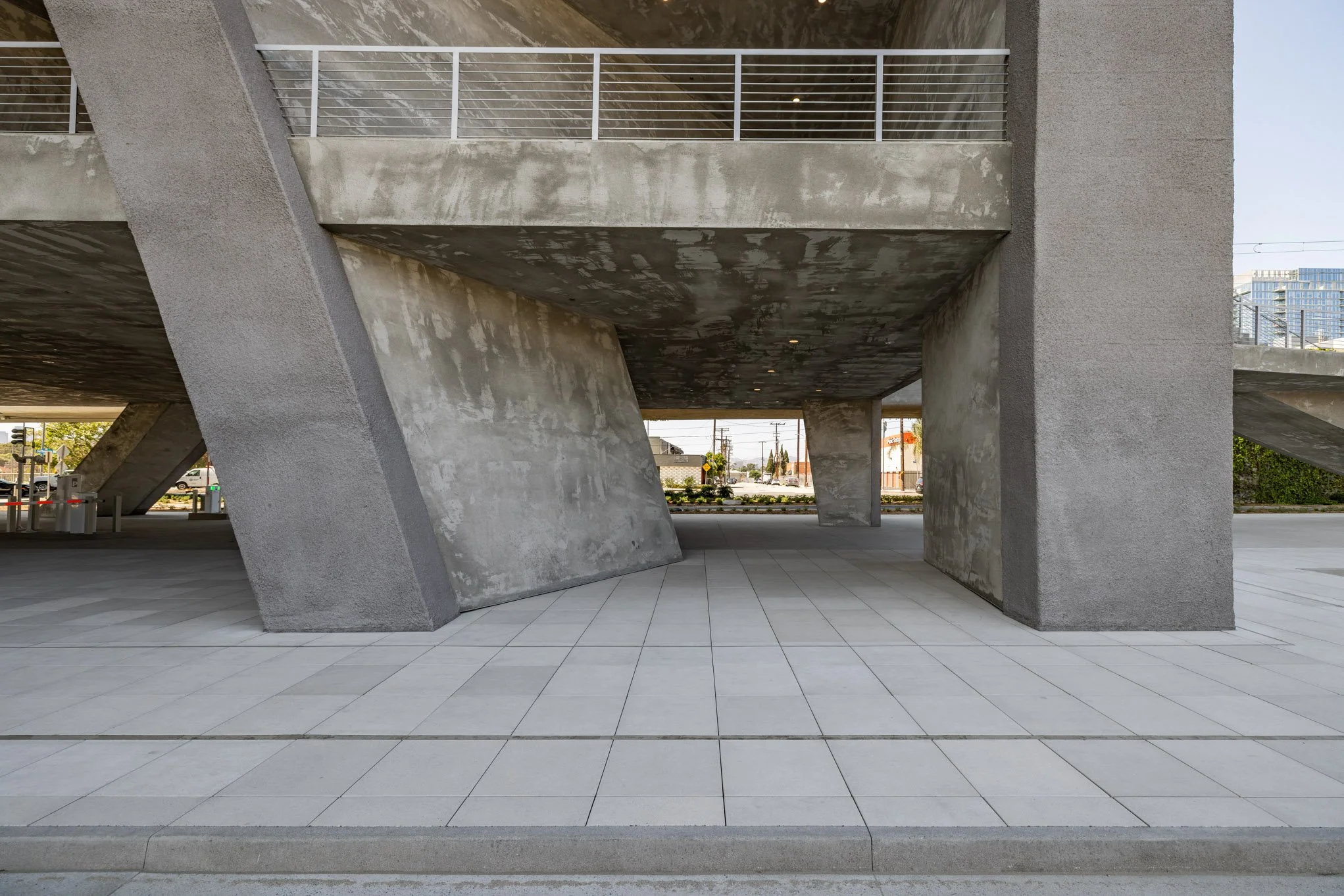 Modern concrete building with geometric architecture, open space underneath elevated structure, tiled sidewalk in foreground.