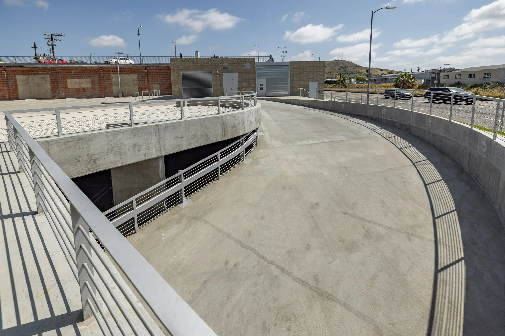Parking structure rooftop with concrete surface, metal railings, and a small building in the background under a partly cloudy sky.