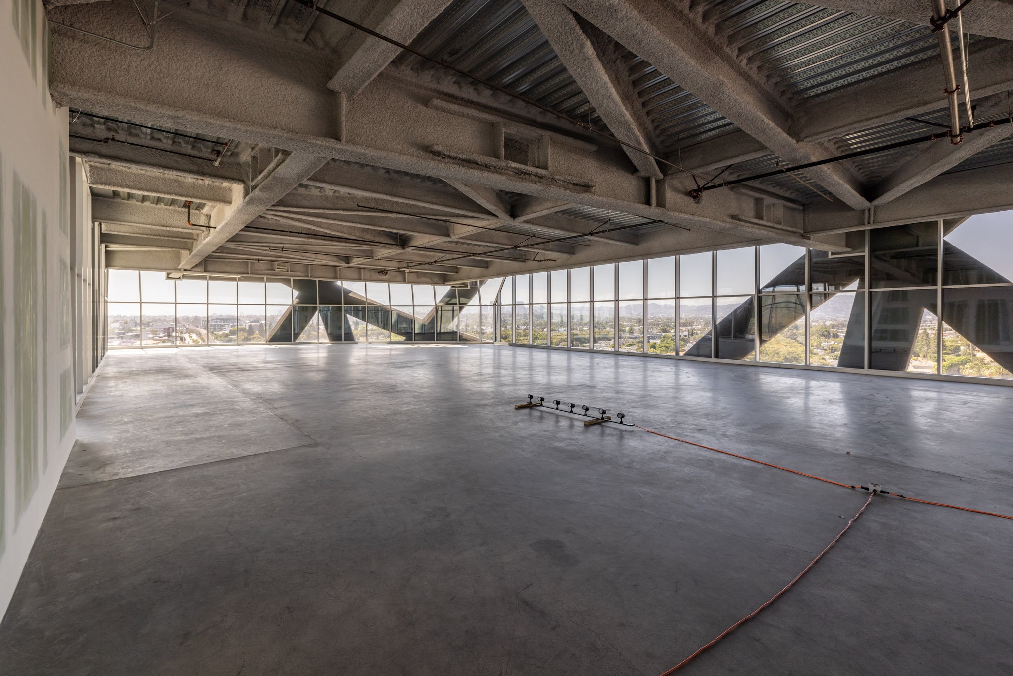 Empty, unfinished high-rise office space with large floor-to-ceiling windows and a view of the city skyline, exposed ceiling beams and ductwork, and construction wiring on the floor.