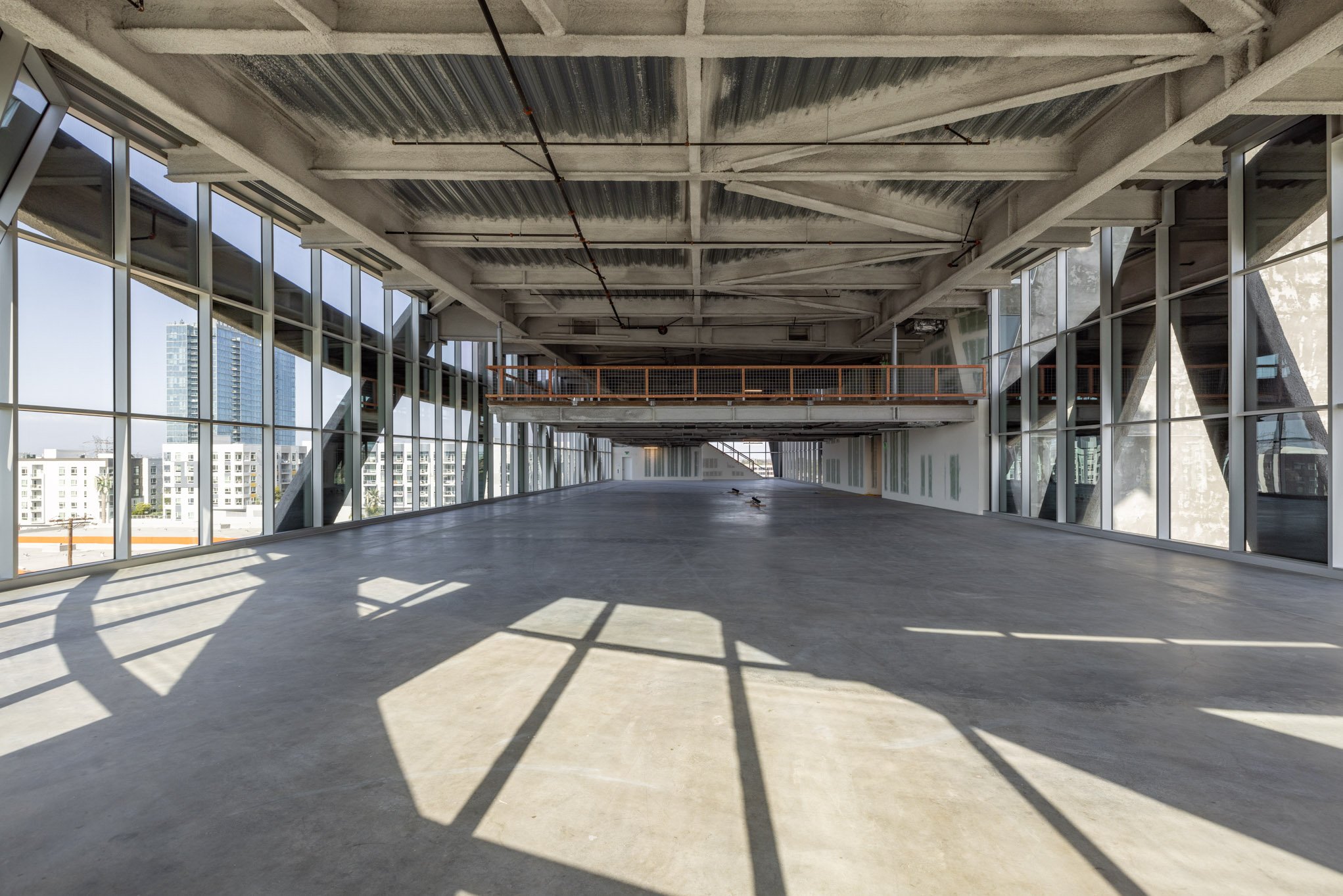 An empty spacious room with large floor-to-ceiling windows, concrete flooring, and an unfinished ceiling with visible beams and pipes. Shadows of window frames cast patterns on the floor.