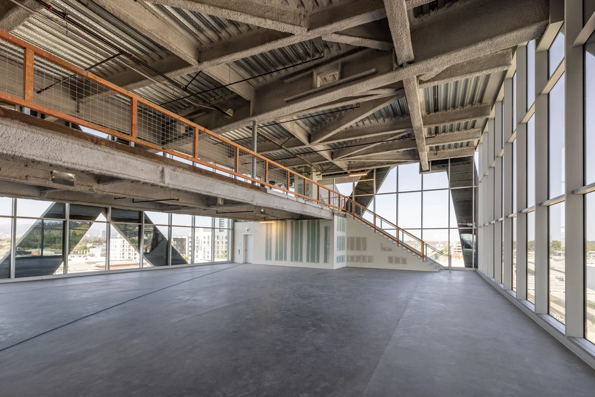 Interior of a modern, unfinished commercial space with large windows, concrete flooring, and an exposed ceiling with beams and insulation. There is a staircase leading to an upper level with a safety railing.