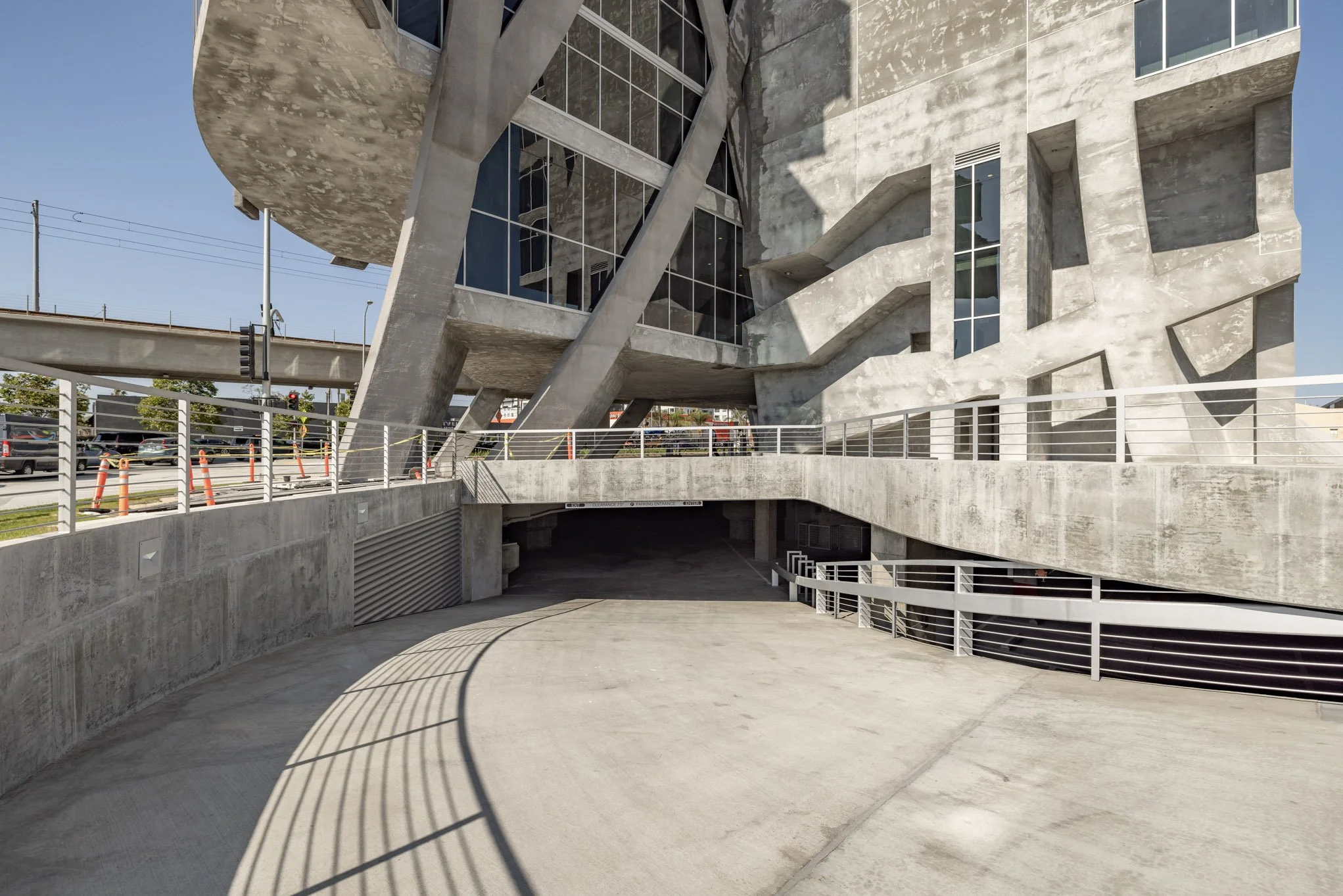 Modern building with concrete facade and angular design, featuring large glass windows, support beams, and a multi-level parking area with concrete walkway and white railings.