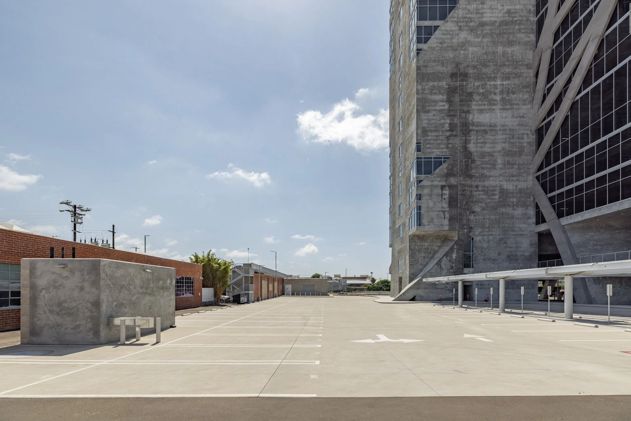 Empty rooftop parking lot with a tall concrete building on the right and smaller brick and concrete structures on the left, under a partly cloudy sky.