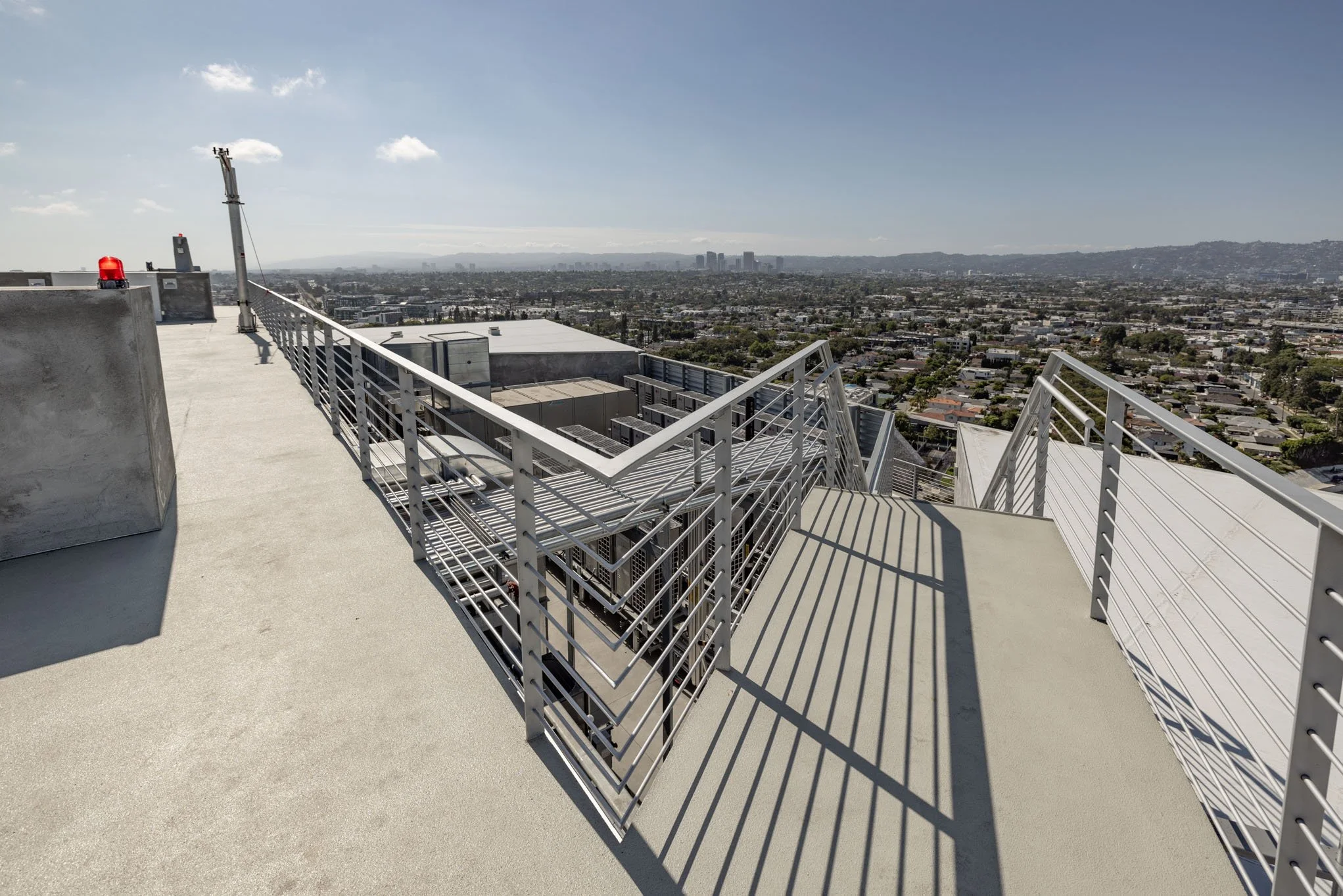 View from the rooftop of a high-rise building showing metal railings, HVAC units below, and a cityscape in the distance under a partly cloudy sky.