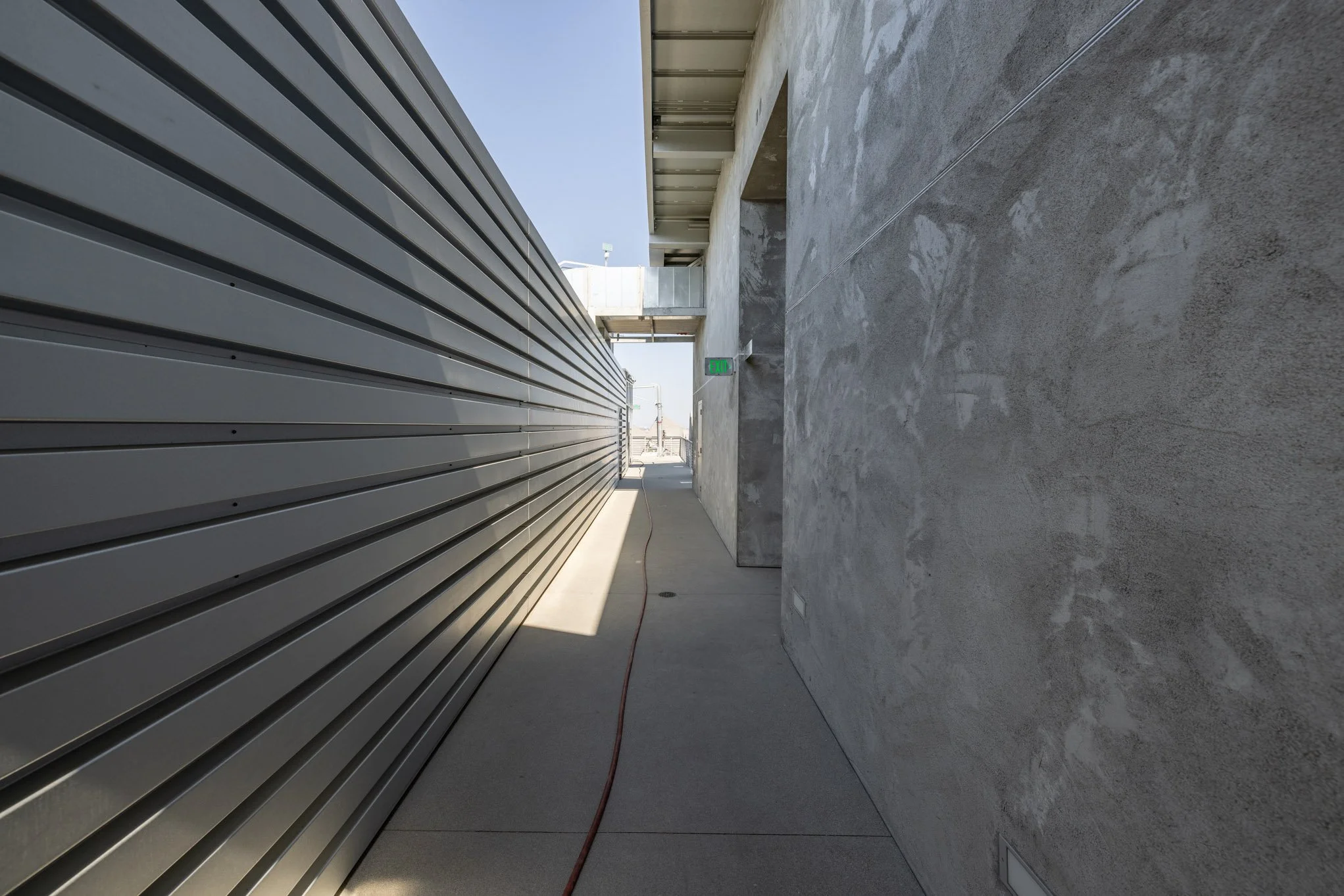 Sidewalk between a metal building and a concrete wall, with an exit sign and a glimpse of the street in the distance.