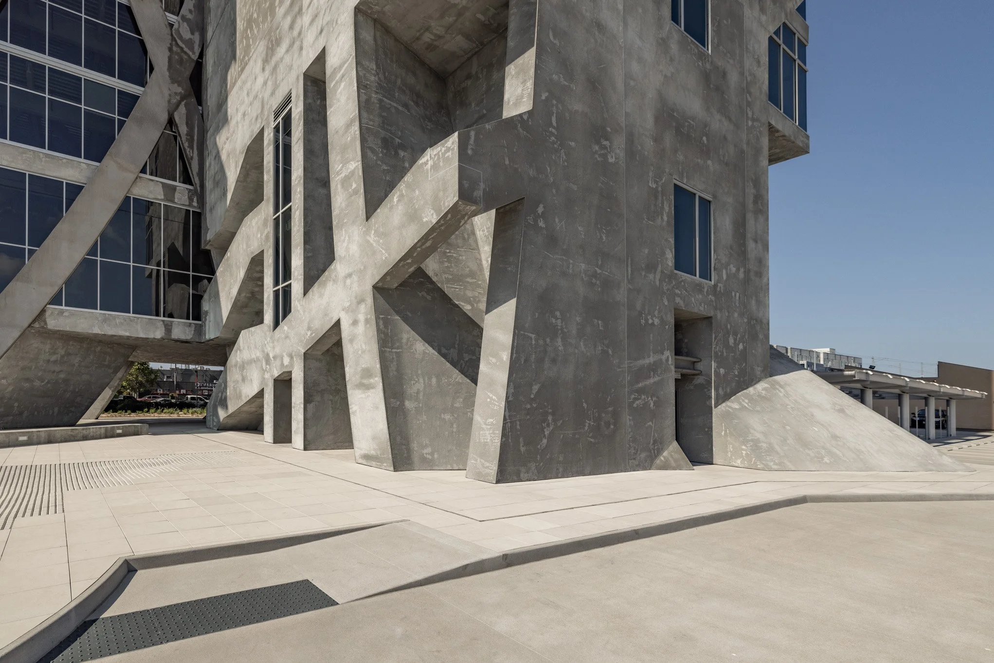 Modern building with concrete and glass architecture, featuring large angular concrete supports and a textured concrete facade under clear blue sky.