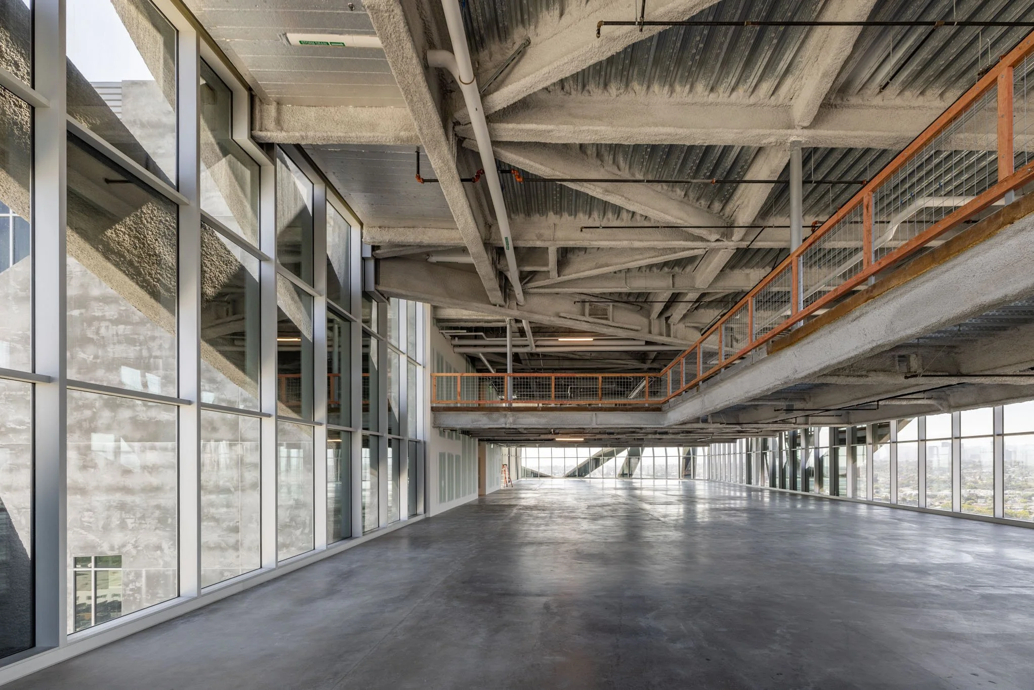 Interior view of a multi-level building under construction with large glass windows, exposed concrete and steel beams, and orange safety railing.