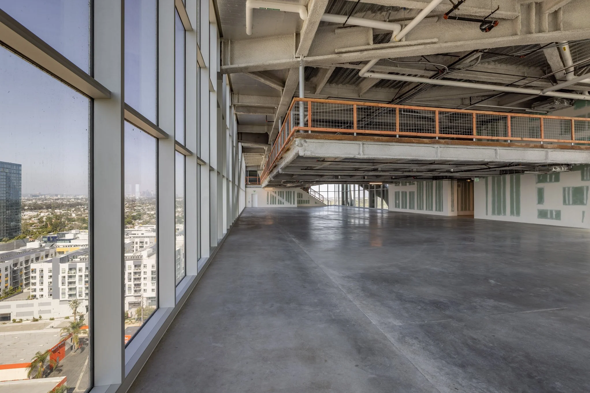 Empty, unfinished interior space with large floor-to-ceiling windows on the left, exposing a cityscape outside, and construction elements like pipes, scaffolding, and partially finished walls.