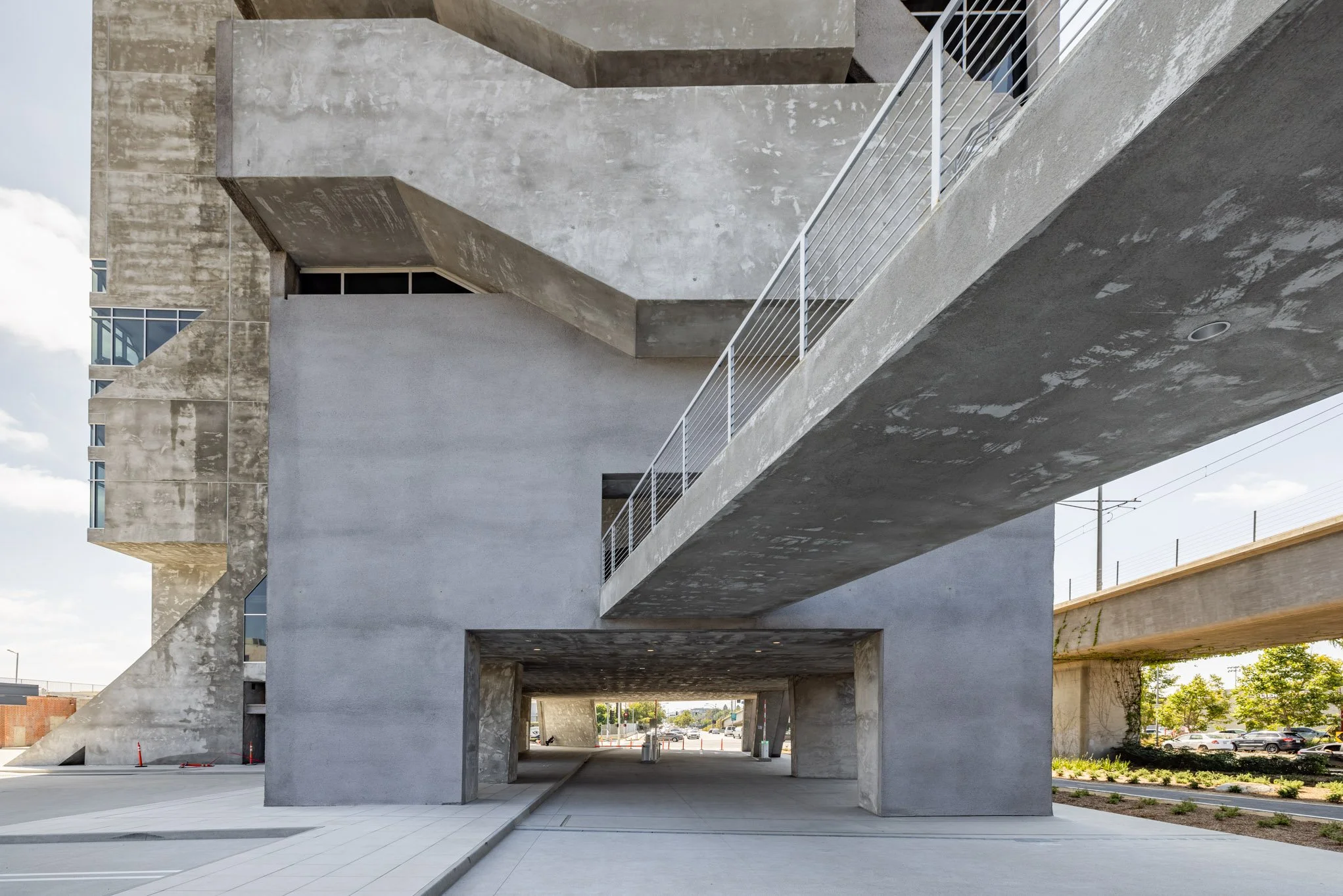 Modern concrete building with geometric design and walkways, underpass visible with a street and trees in the background.