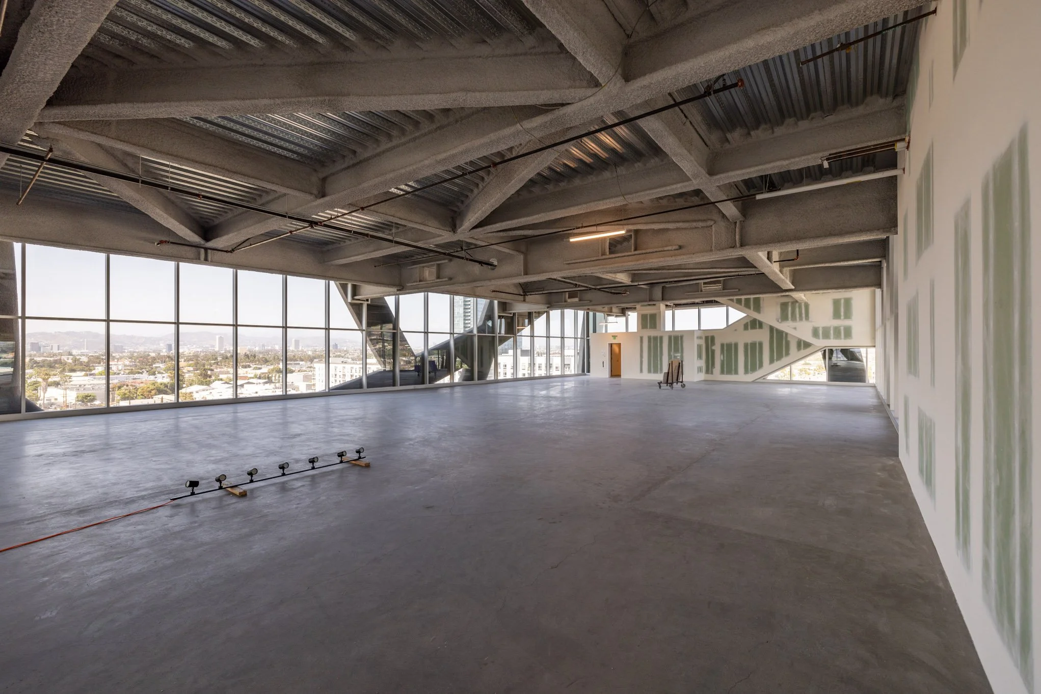 Empty modern interior space with large floor-to-ceiling windows, unfinished walls, and visible ceiling structures, with cityscape view outside.