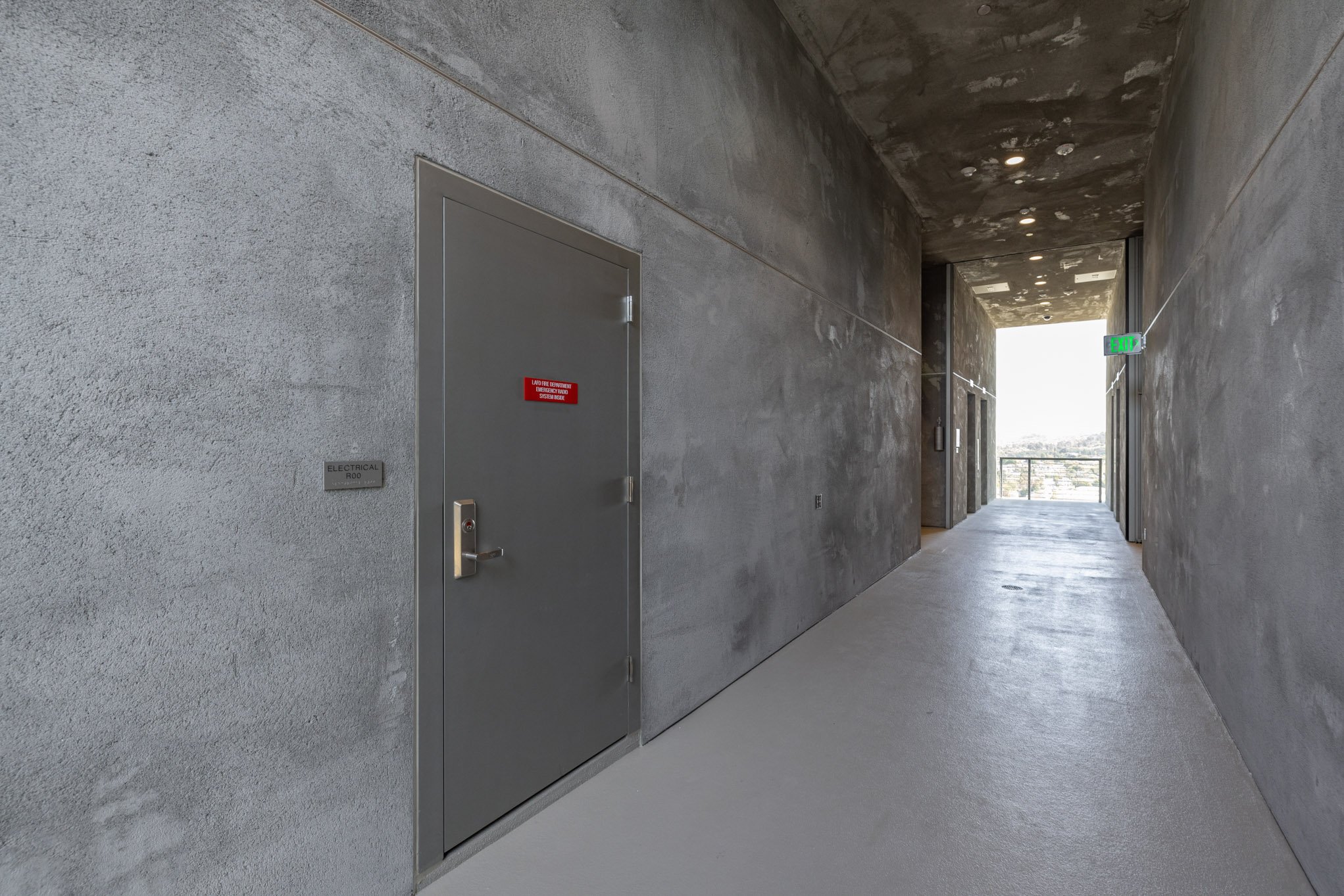 Concrete hallway with gray walls, a gray door labeled 'Electrical R00', and an illuminated exit sign, leading to an outdoor view.
