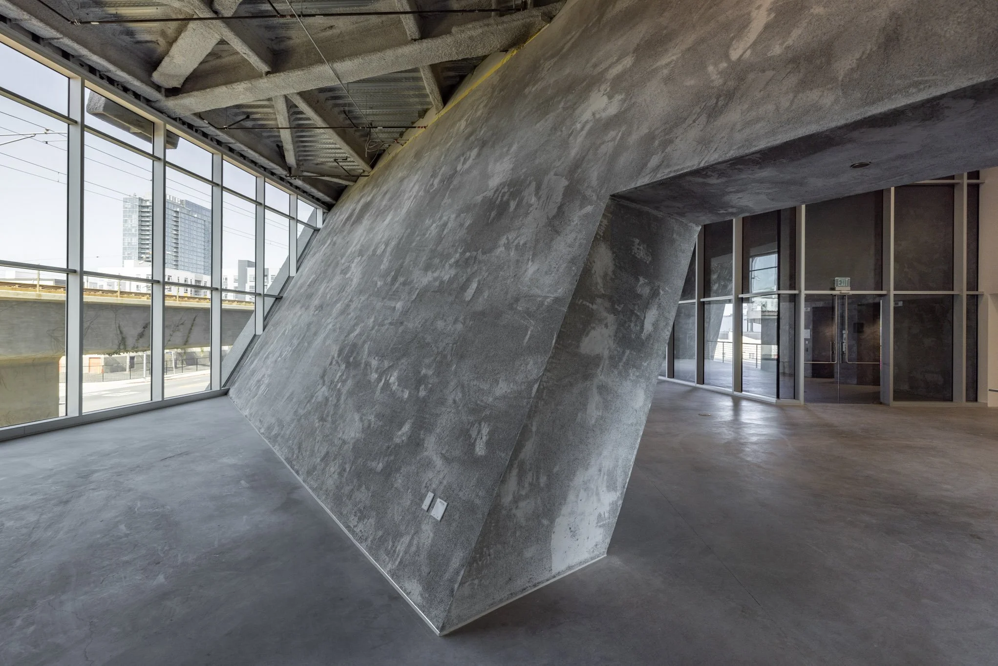 Modern interior space with a large, angular, concrete wall feature, floor-to-ceiling windows, and exposed structural elements.