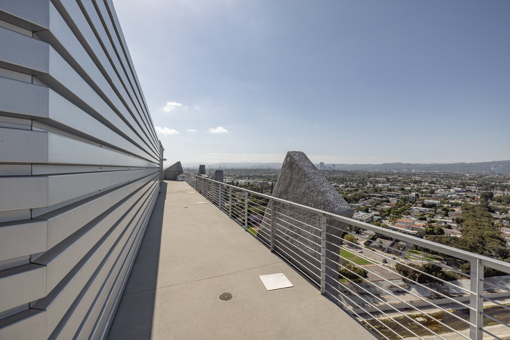 View from a rooftop balcony with metal railing, overlooking a cityscape with a unique angular building and mountains in the distance.