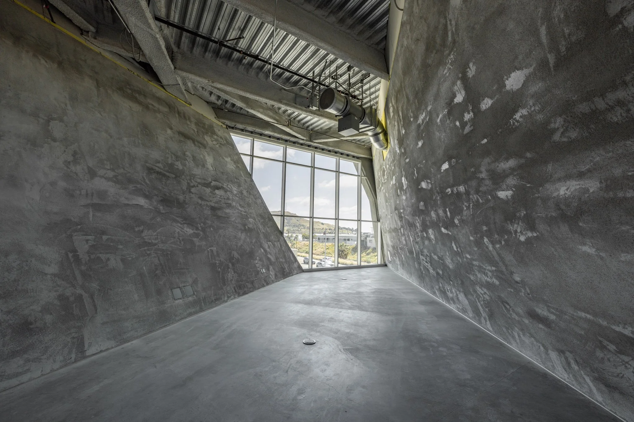 Empty, unfinished room with large window, concrete walls, ceiling with visible ductwork and wiring, and smooth concrete floor.