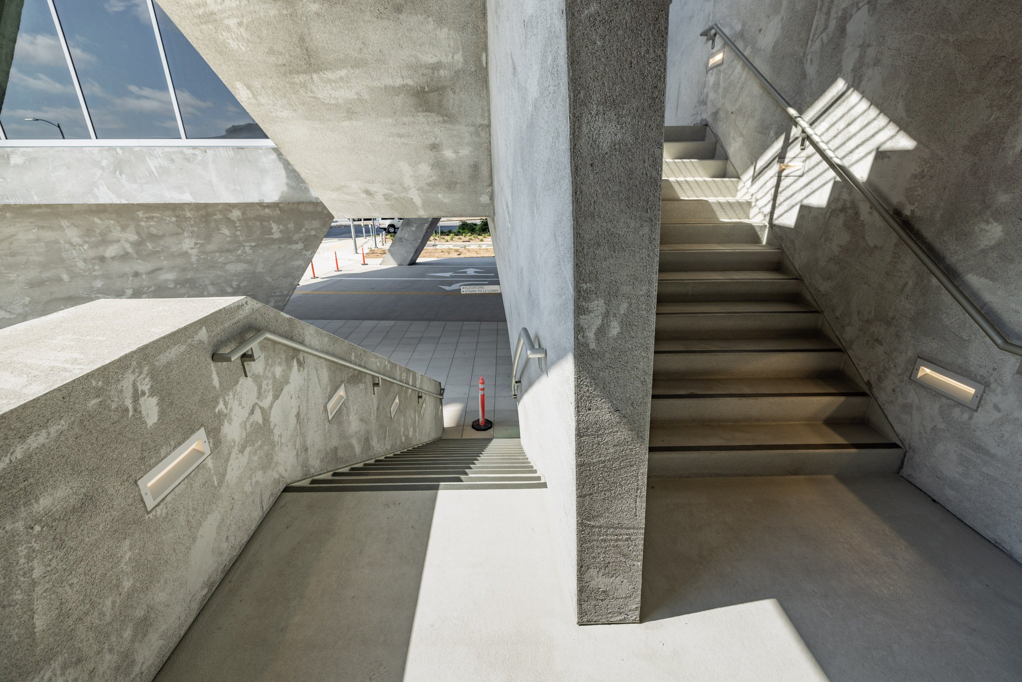 Concrete outdoor staircase with metal handrails, sunlight casting shadows, part of a modern building with large glass windows.