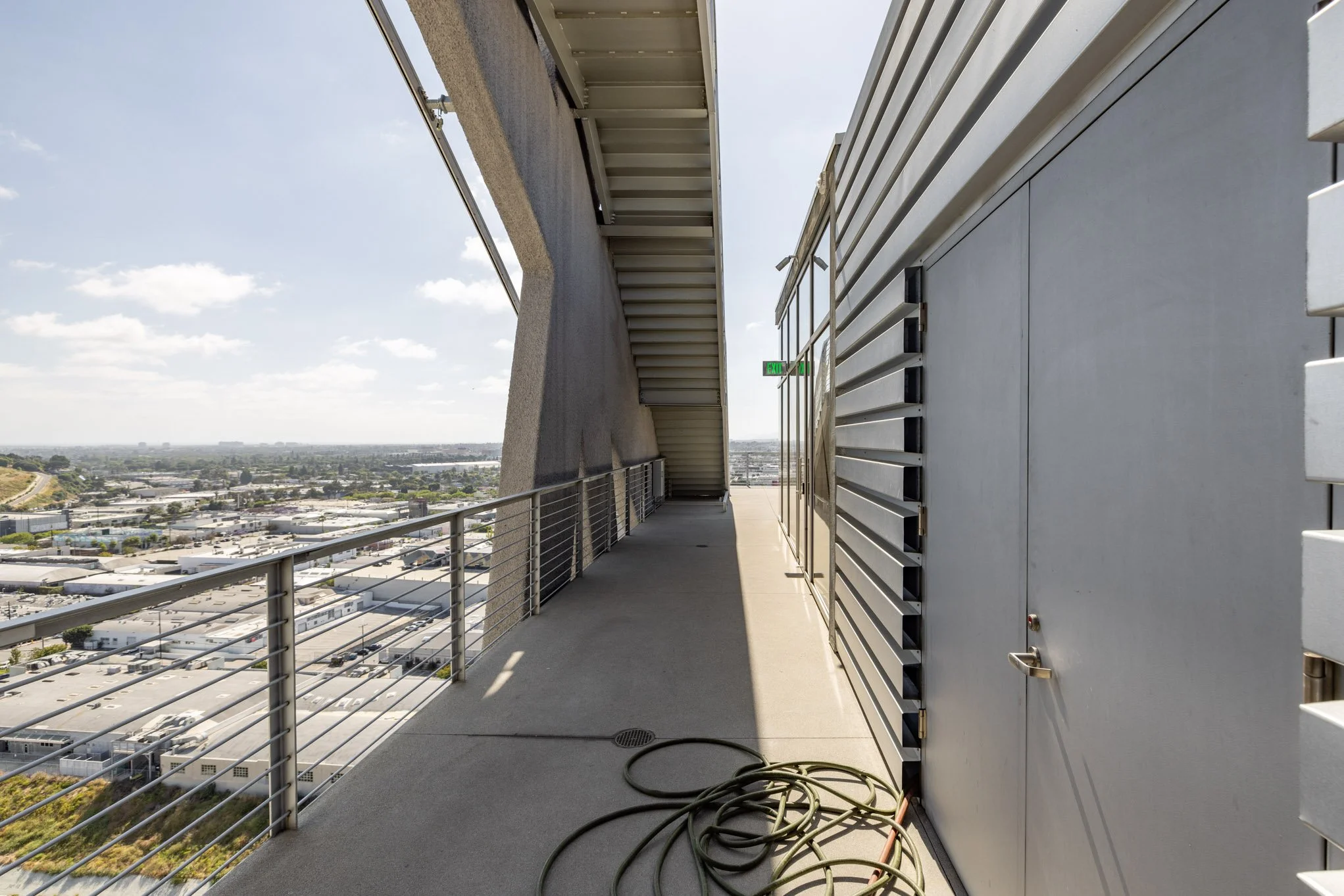 An empty rooftop balcony with a metal railing, outdoor electrical cable, and view of a cityscape and sky in the background.