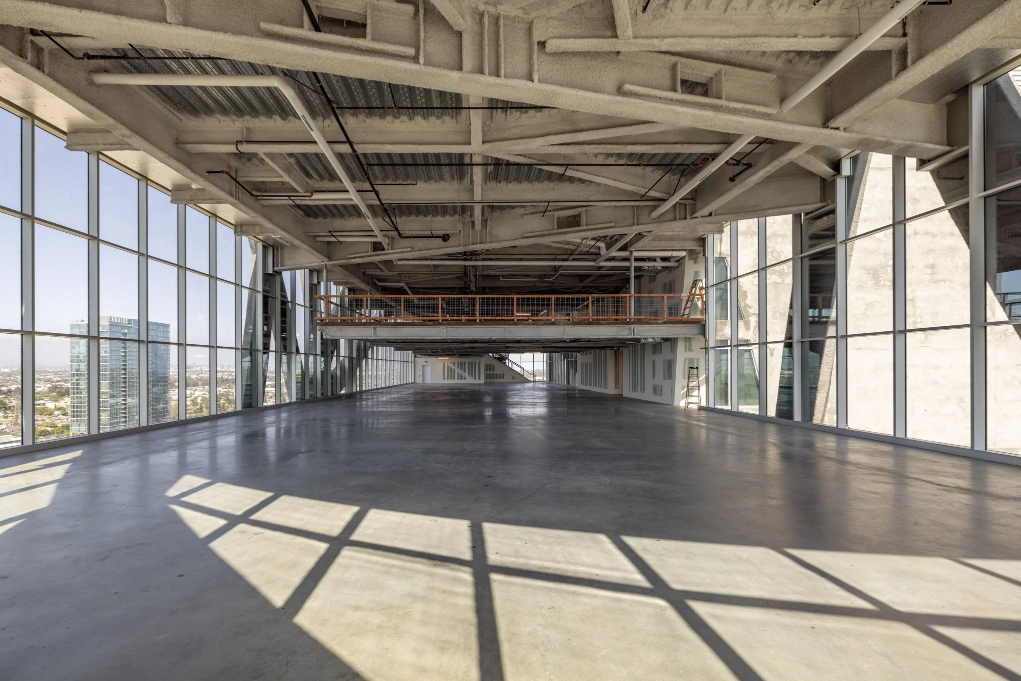 Empty interior space of a modern building with large floor-to-ceiling windows, concrete floor, exposed ceiling with piping, and a suspended balcony or mezzanine level.