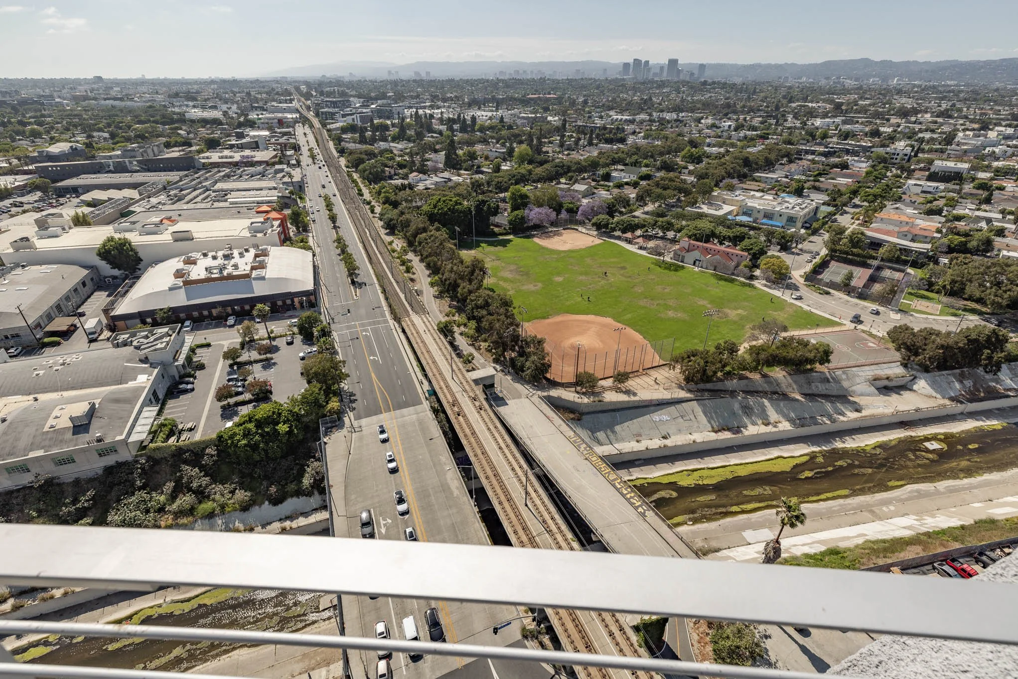 Aerial view of an urban area showing a street, railway tracks, a large park with tennis courts and baseball field, residential neighborhood, and city skyline in the distance.