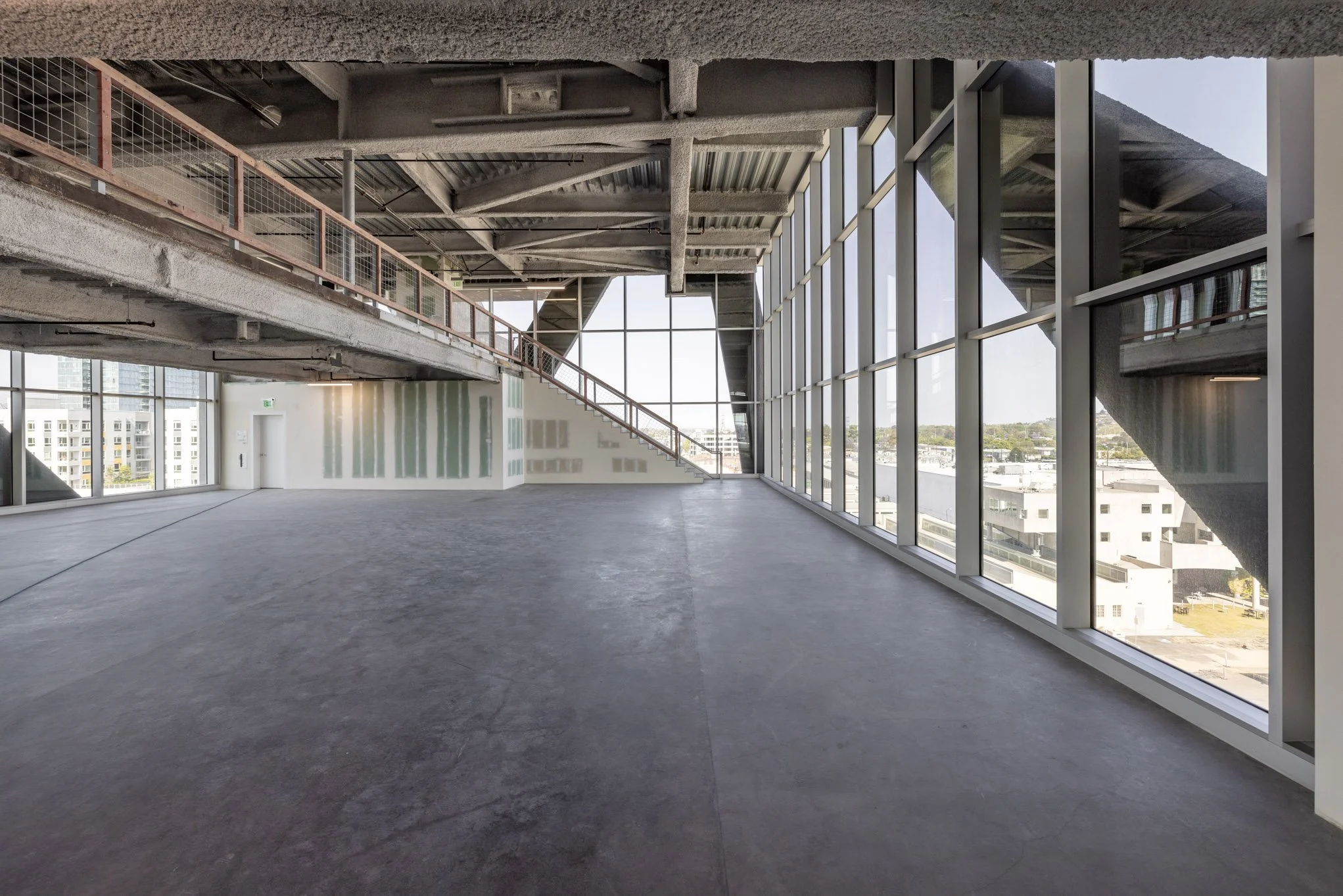 Empty interior space with large floor-to-ceiling windows, concrete floors, and exposed ceiling beams, part of a modern building under construction or renovation.