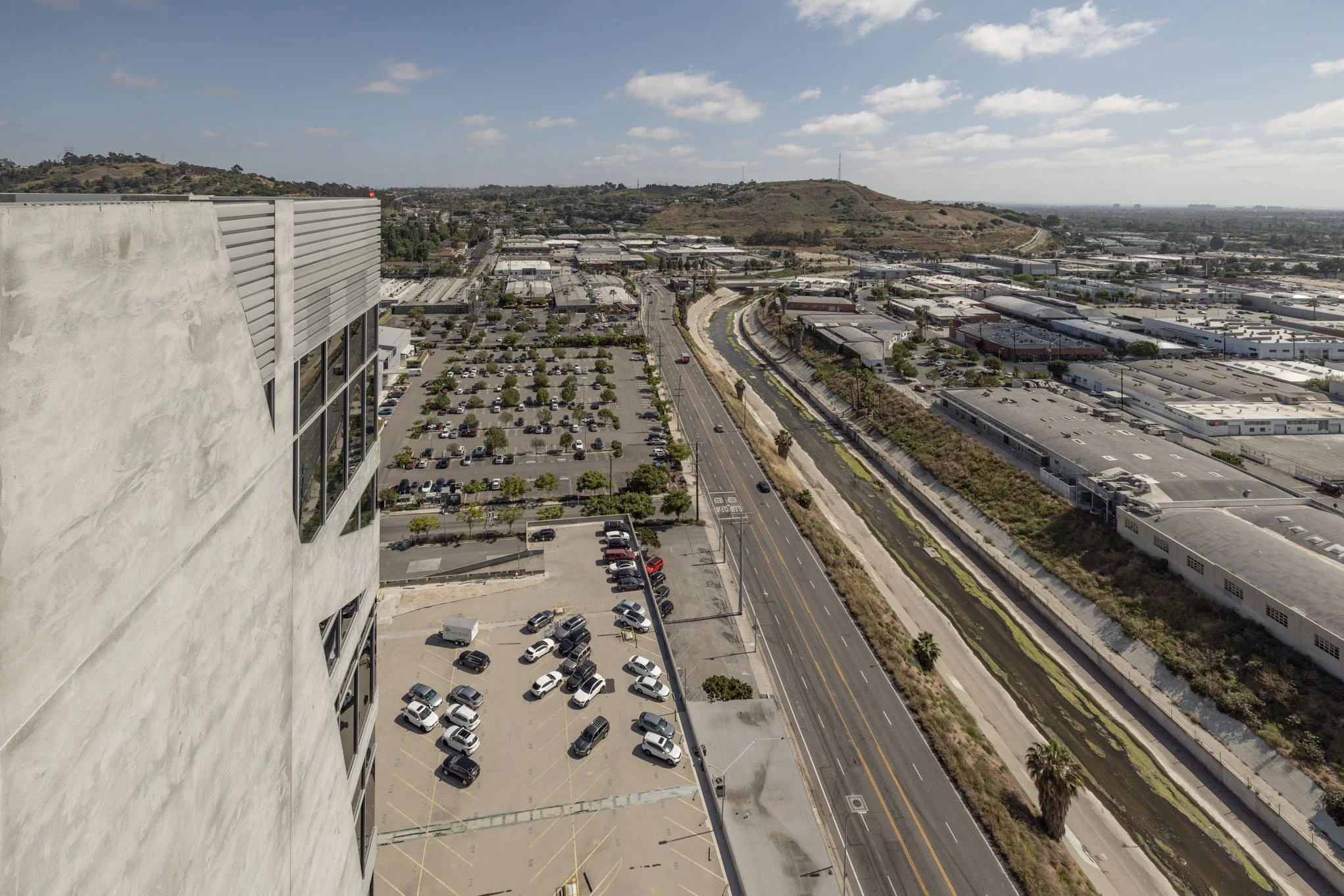 Aerial view of a cityscape with a large parking lot, a road with few cars, industrial buildings, and a hill in the background under a partly cloudy sky.