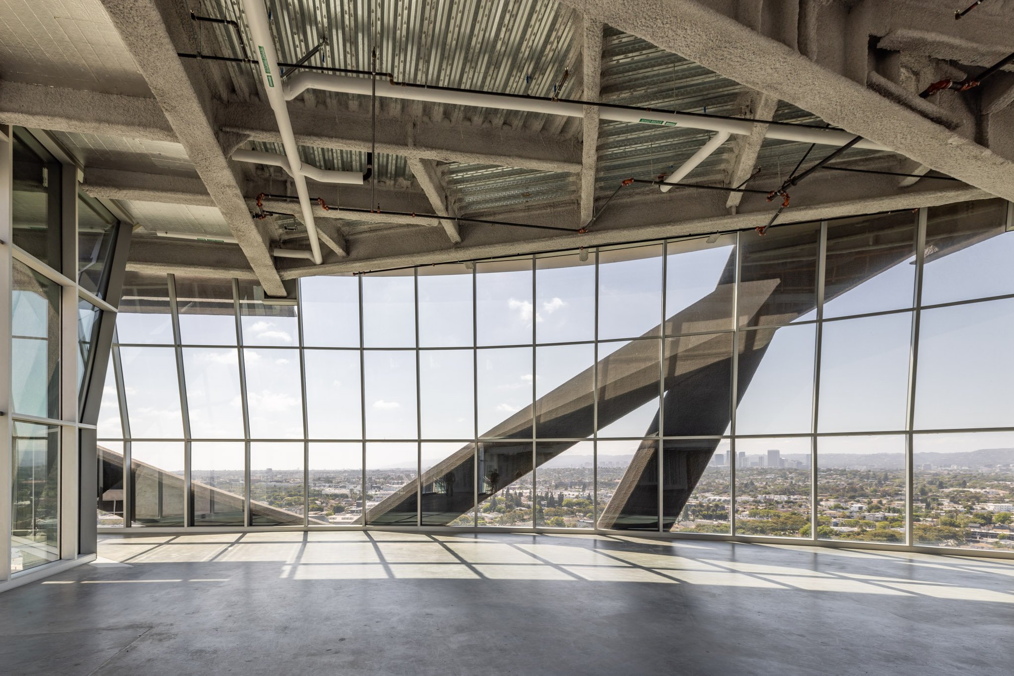 Empty room with floor-to-ceiling windows overlooking a cityscape, exposed concrete ceiling with pipes and beams, and a concrete floor.