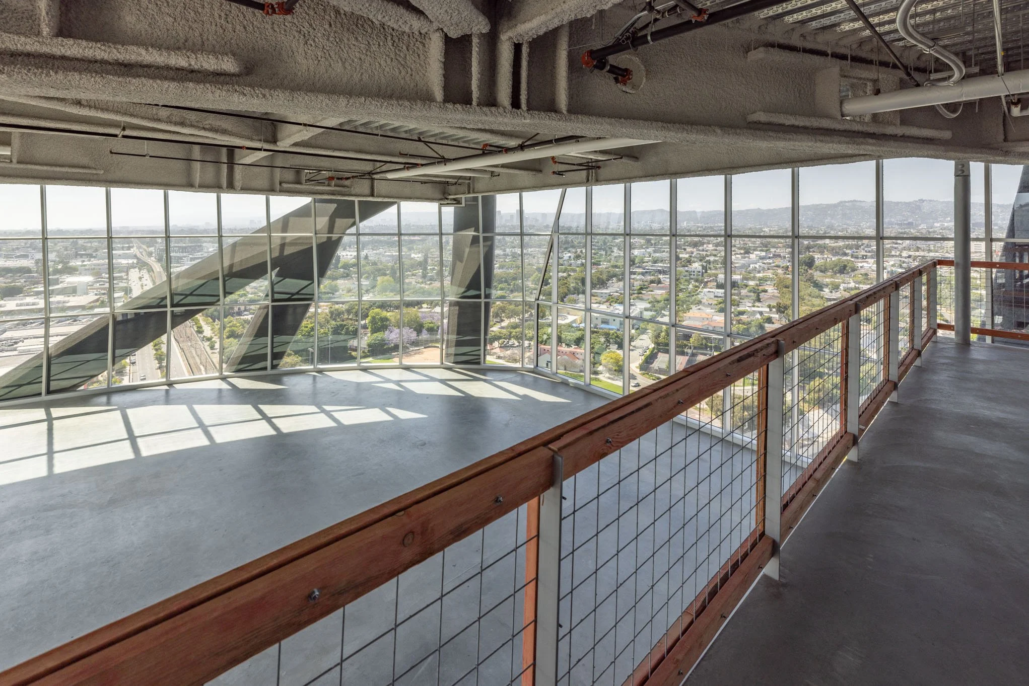 Empty indoor observation deck with wooden railing, large floor-to-ceiling windows, and cityscape view, including residential neighborhoods and distant hills.