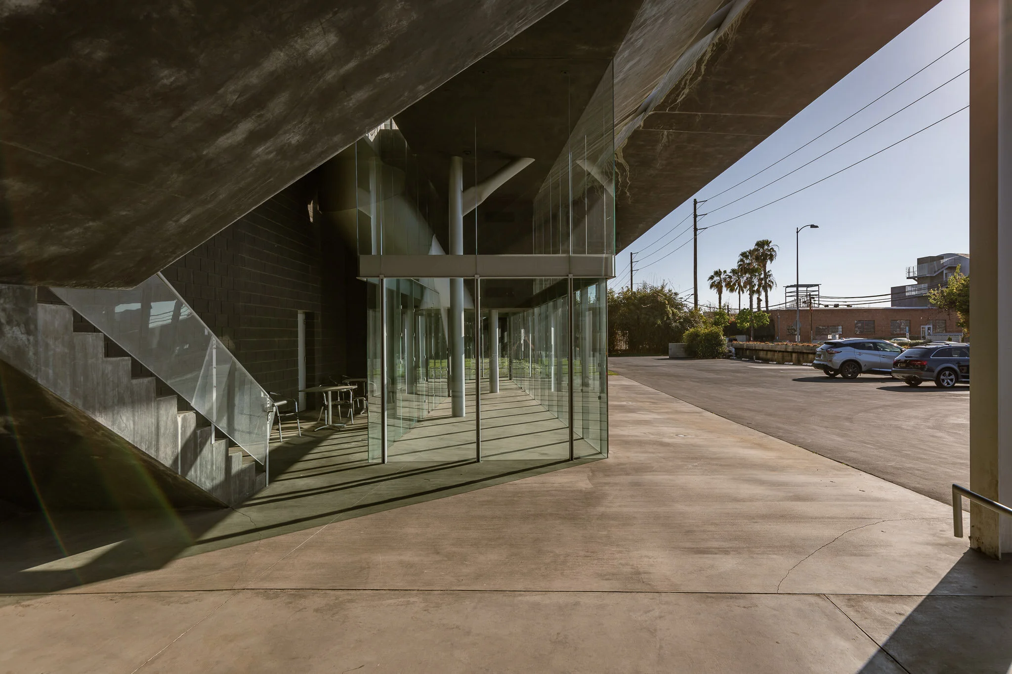 Modern building with glass walls casting shadows on the sidewalk outside on a sunny day, with parked cars and palm trees in the background.