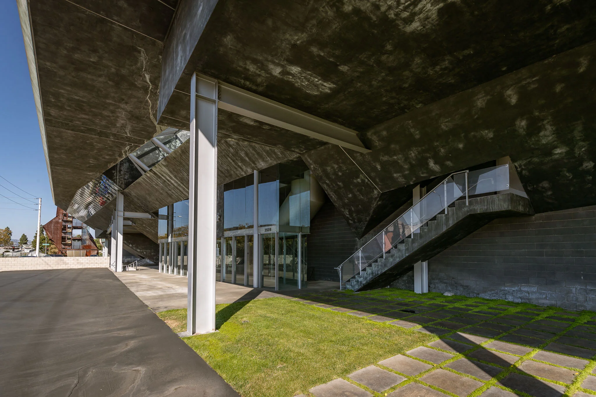Modern building with large glass doors, concrete stairs with glass railings, and an overhanging concrete roof, with a grassy and paved area in front.