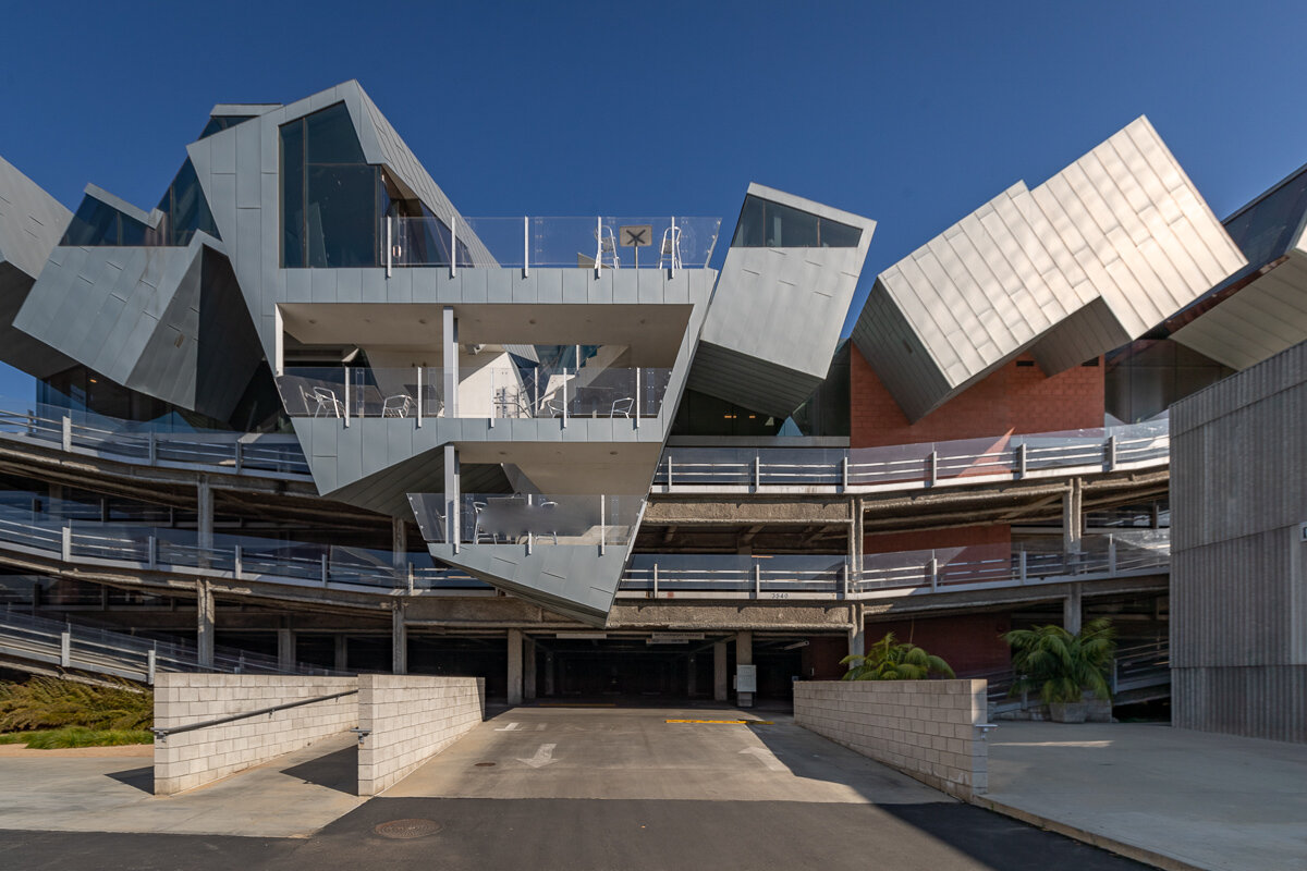 Modern architectural building with unusual angular design, multiple levels, and metallic exterior against a blue sky.