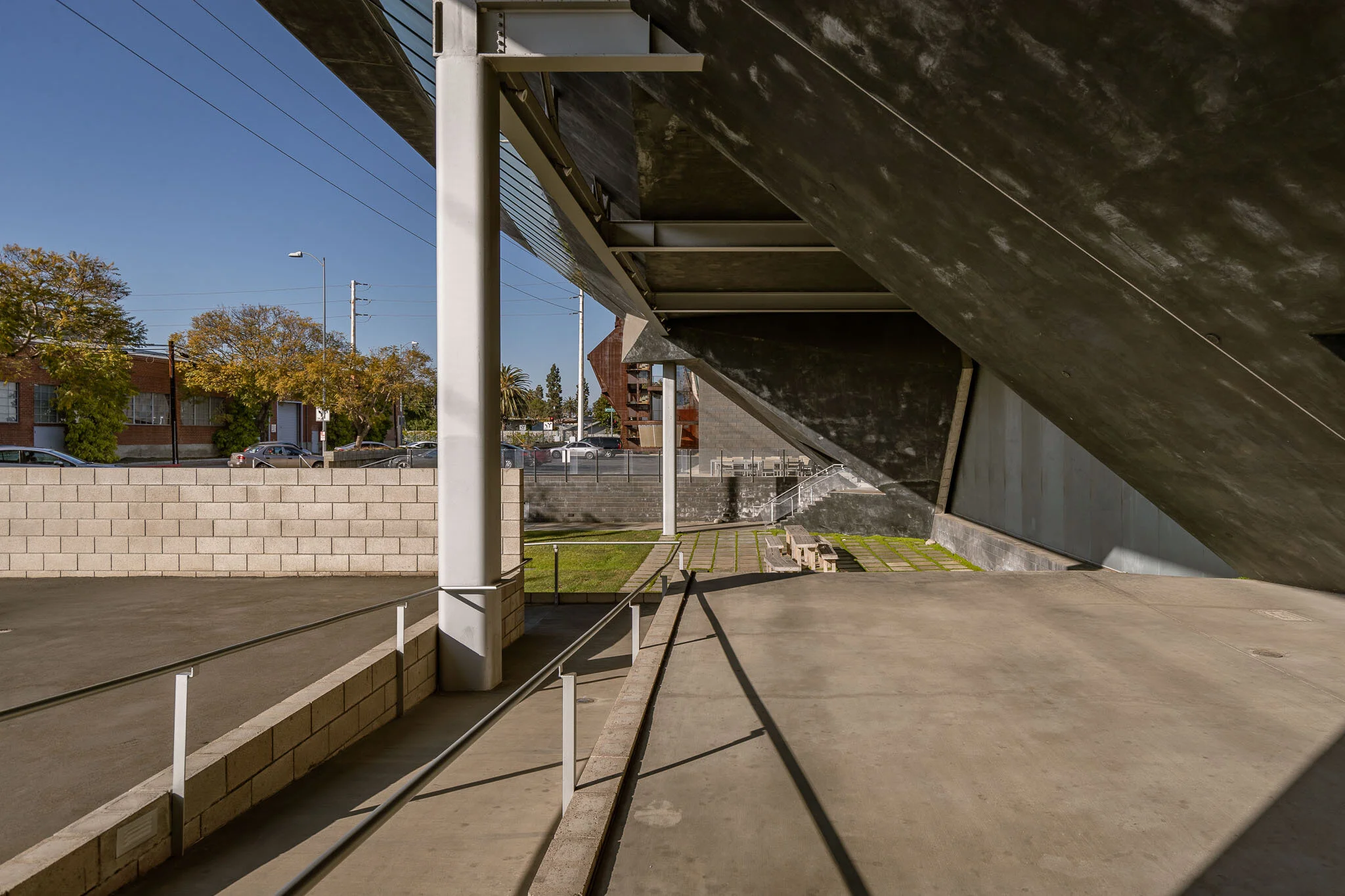 Empty outdoor space with concrete flooring and a large slanted dark wall, some small benches, a grassy area, and a brick wall in the background. Trees and parked cars are visible beyond the wall, with a clear sky overhead.