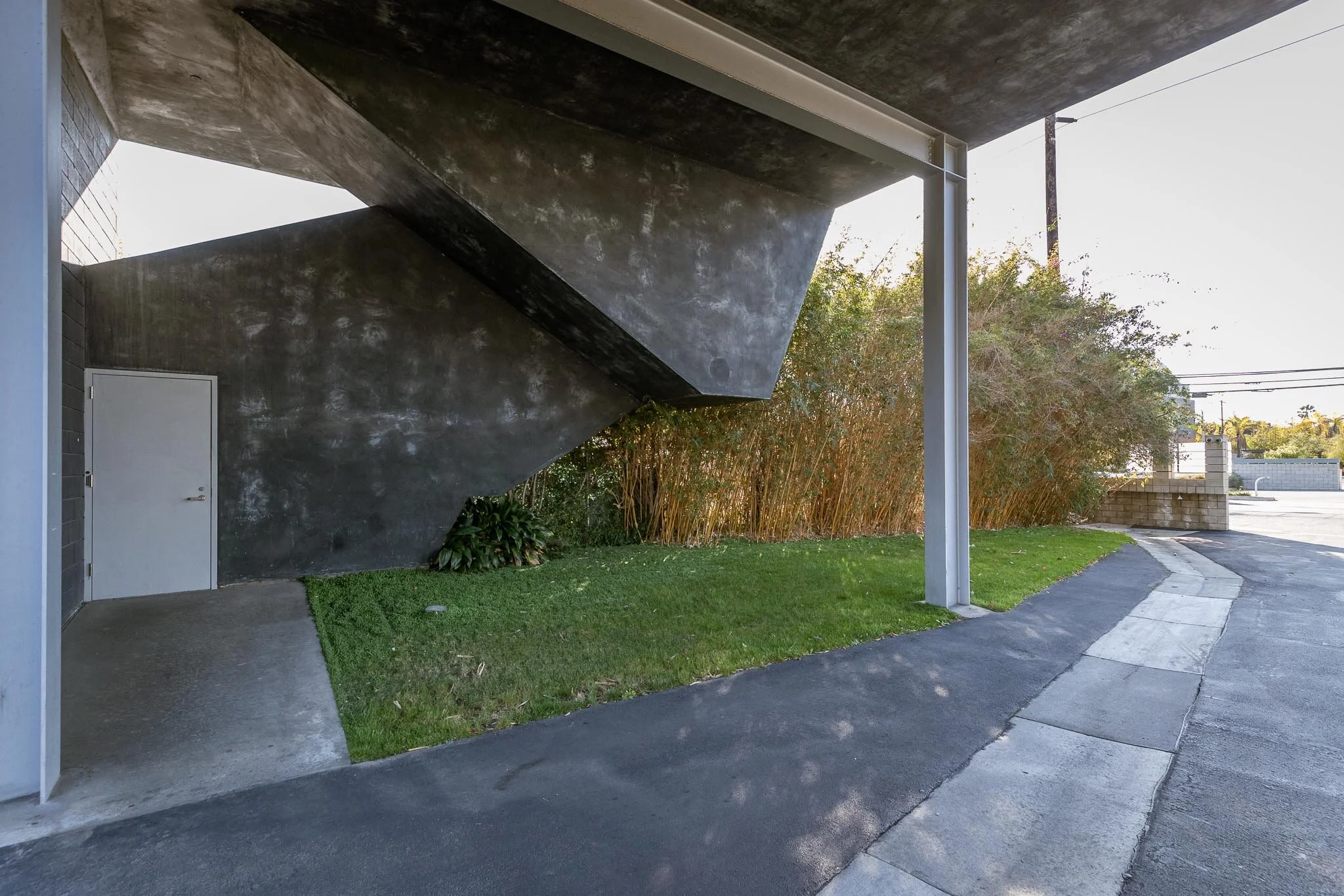 Modern building with a large black concrete overhang, small grass patch, and bushes, next to a sidewalk and street in a sunny area.