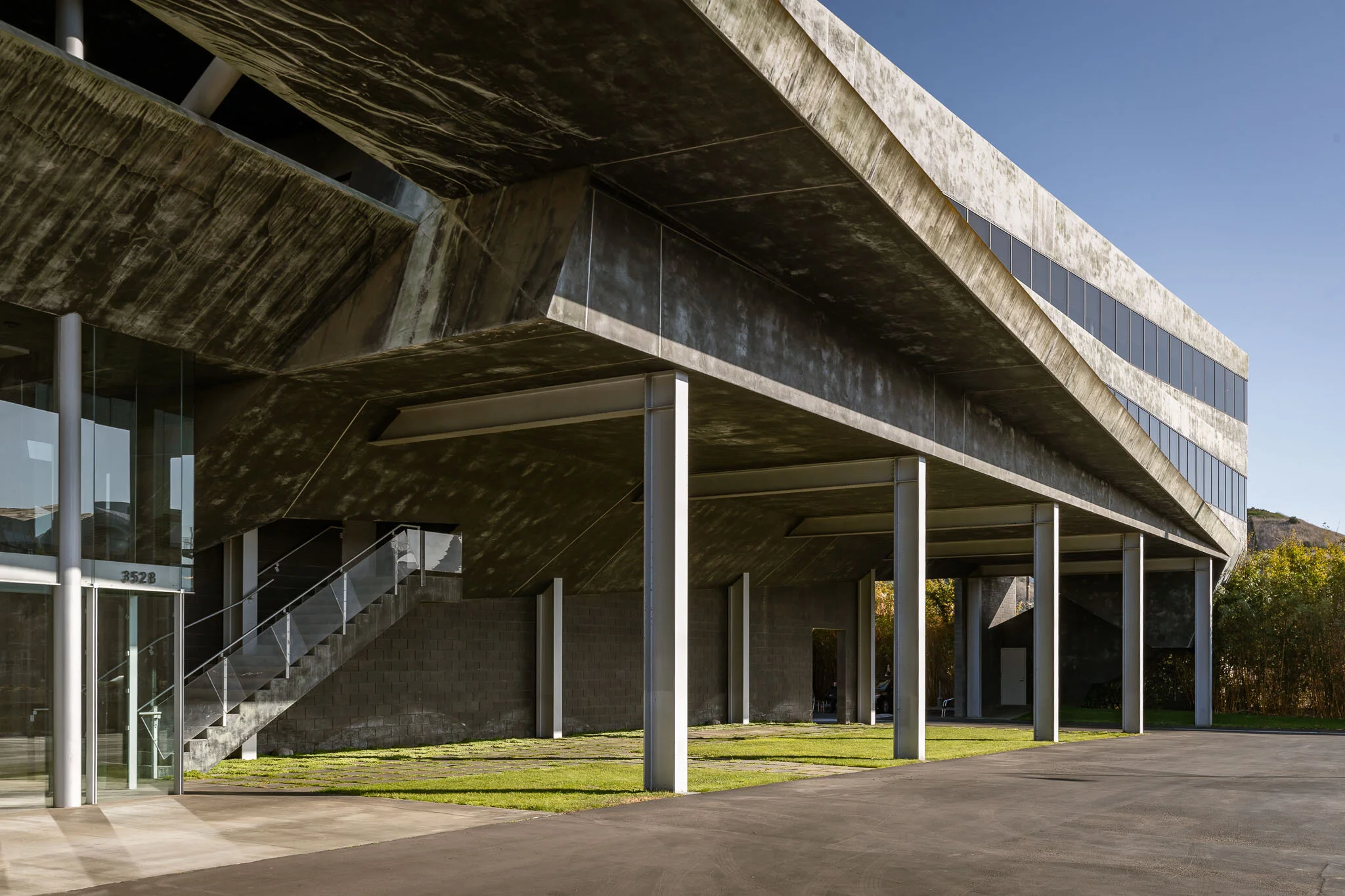 Modern concrete building with glass windows, supported by metal columns, with a staircase leading to the upper level, and a grassy area and driveway in the foreground.